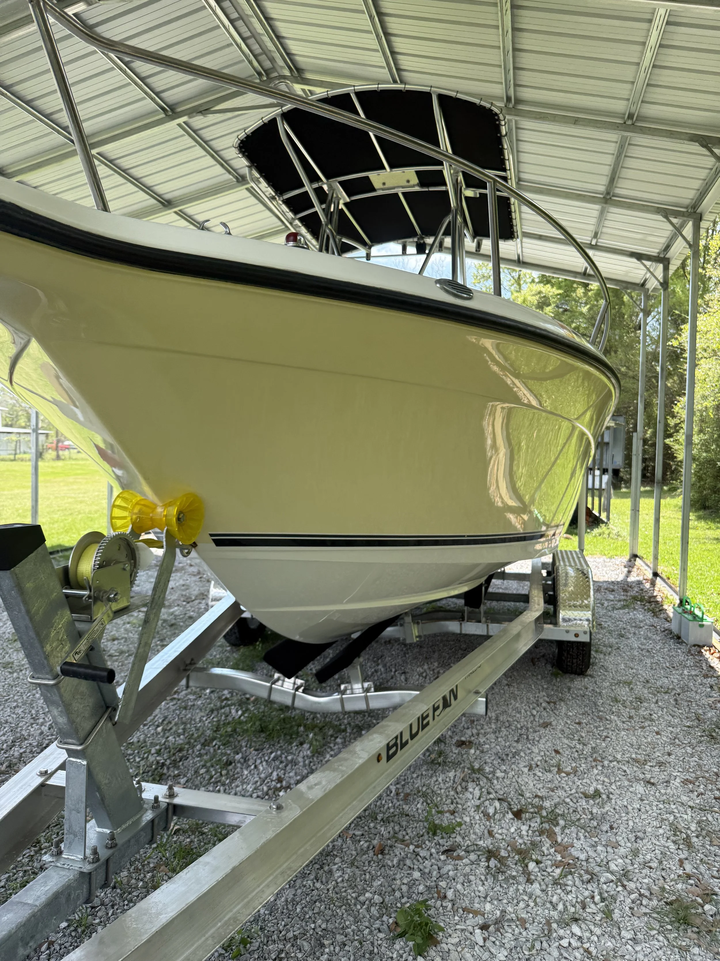 A yellow and white motorboat on a trailer, parked under a metal roof shelter with trees in the background.