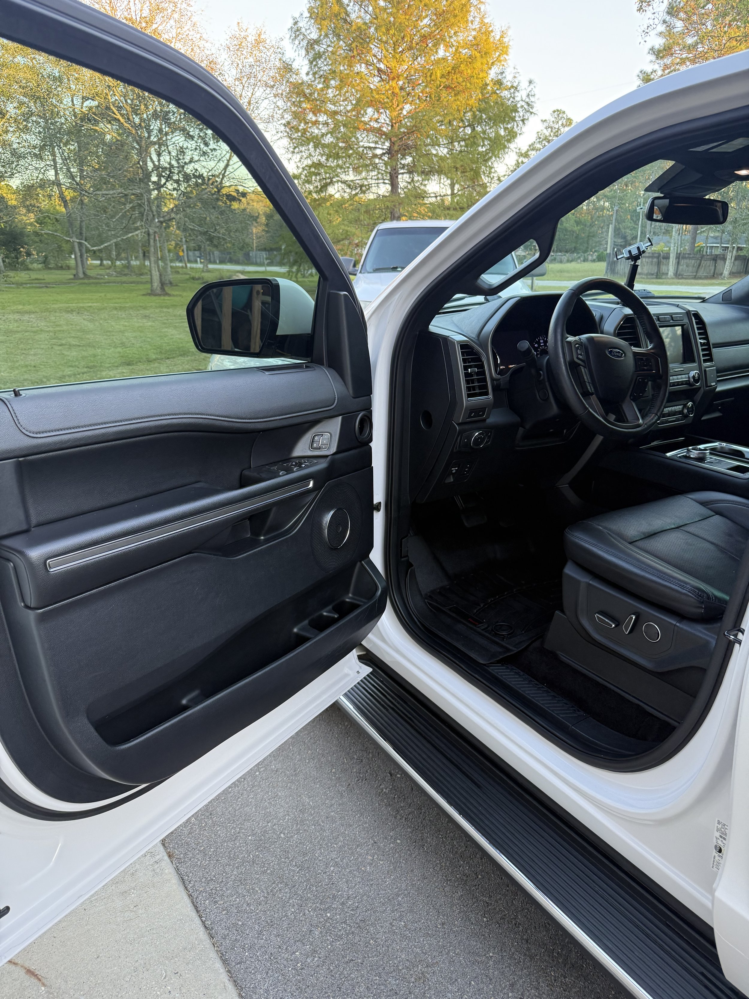 Open car door showing the interior of a black vehicle with a view of the dashboard, steering wheel, and front seat, parked on a driveway with trees and grass in the background.