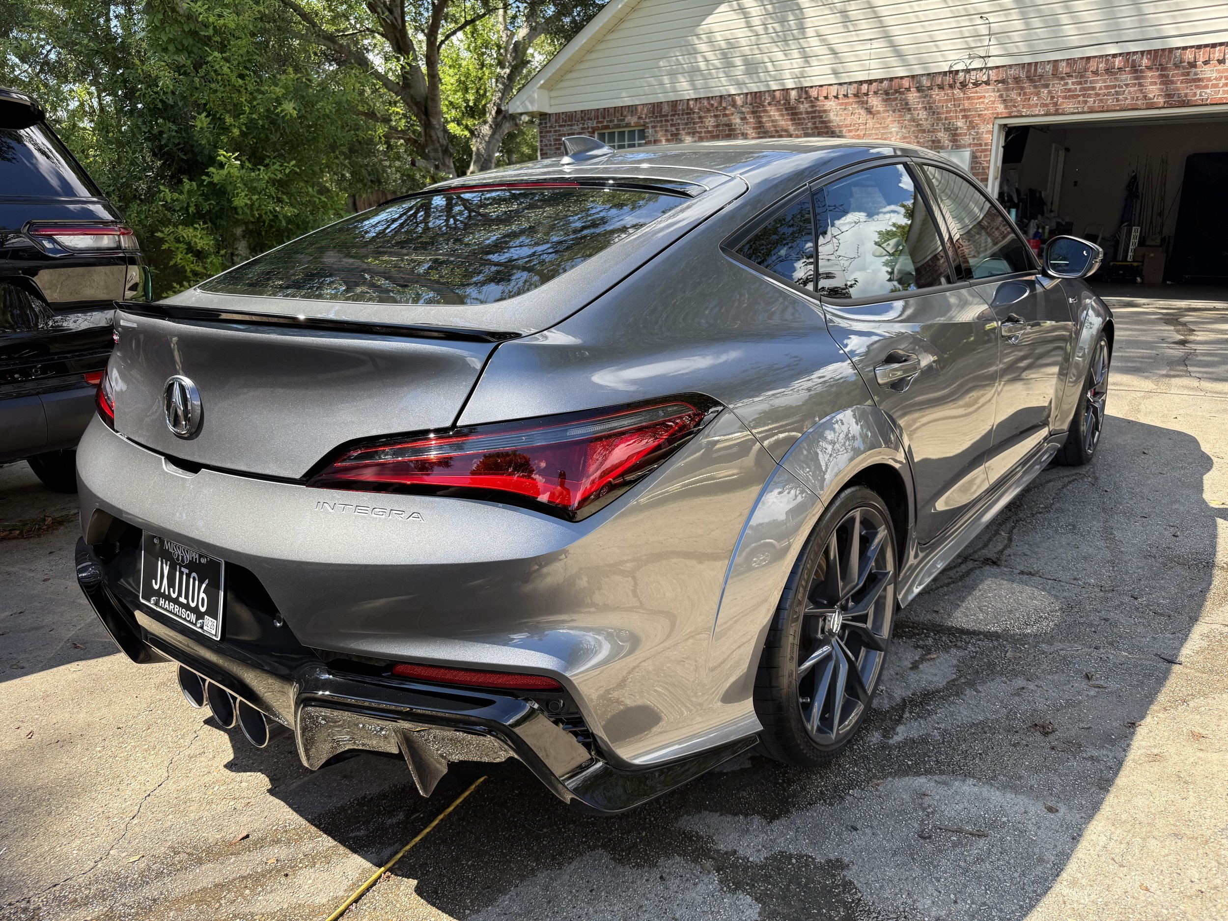 Silver Acura Integra sports car parked in driveway near a garage, with a black vehicle visible in the background, trees, and a house with a brick and siding exterior.