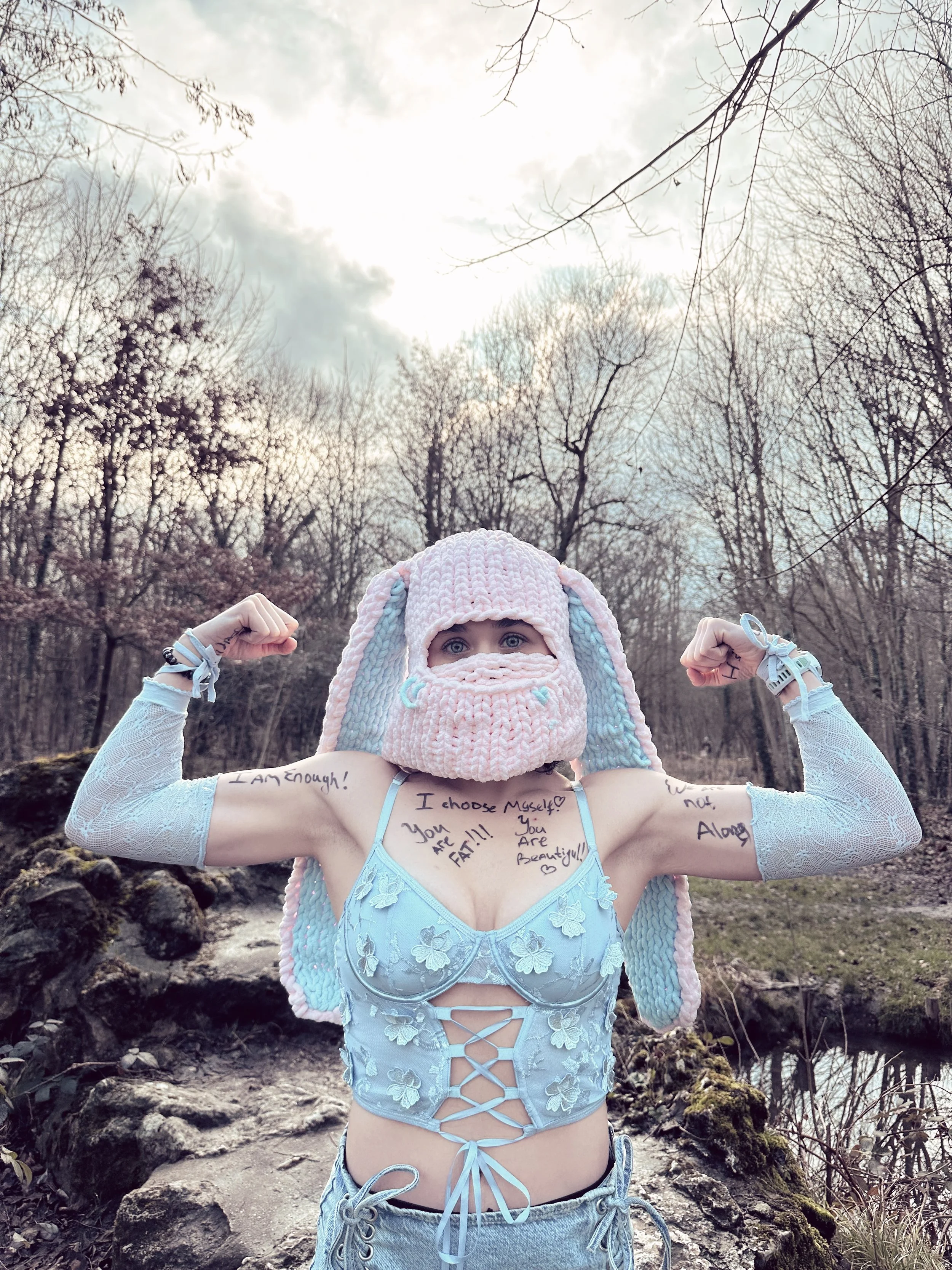 A woman in a pink and blue bunny hat, lace arm warmers, and a lace-up crop top, standing outdoors by a stream with trees and clouds in the background. She is flexing her muscles and has written messages on her arms and chest, expressing self-acceptance and confidence.