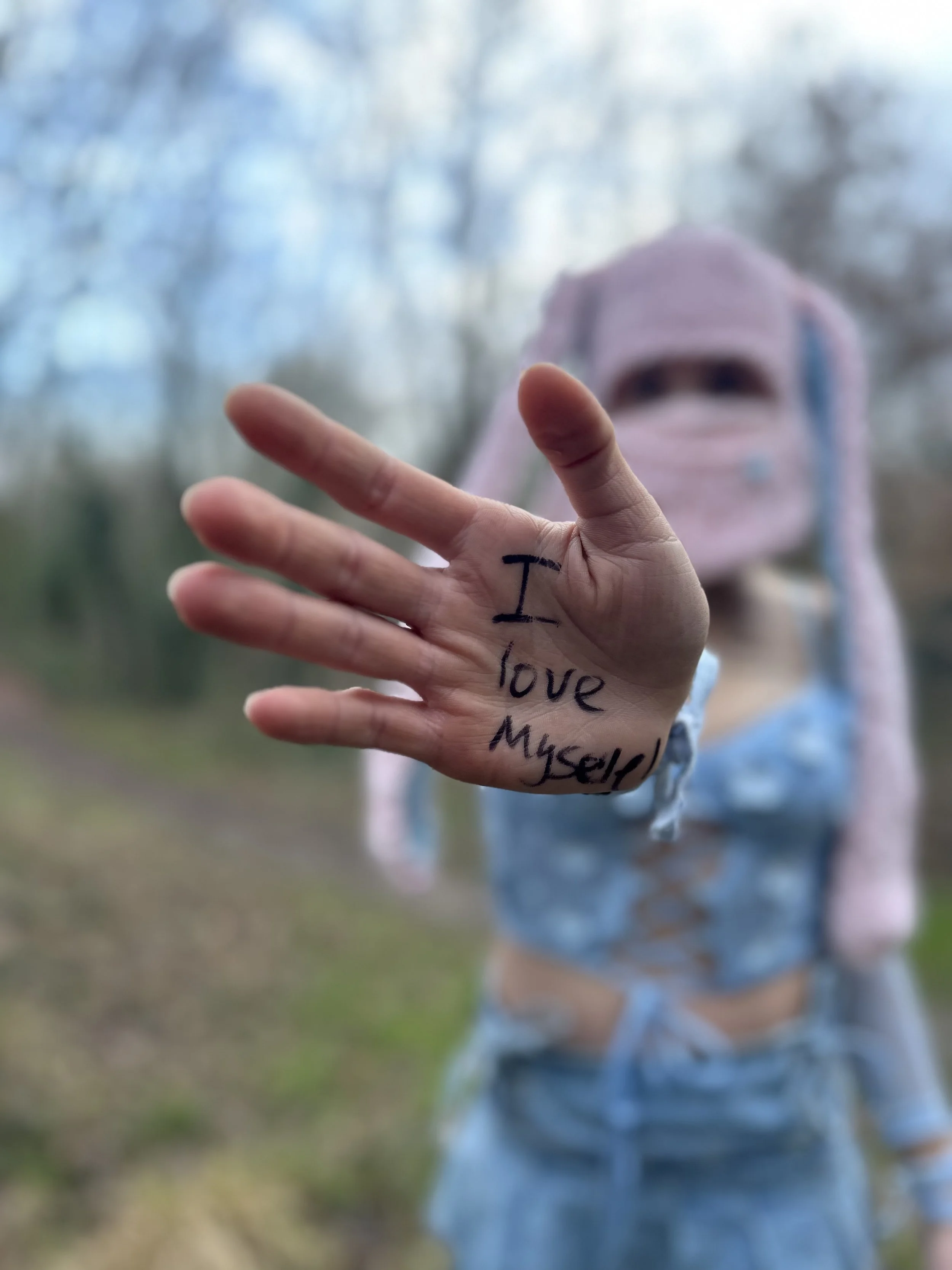 A person with a pink hooded jacket holding out their hand with the words 'I love myself' written on their palm, blurred trees in the background.