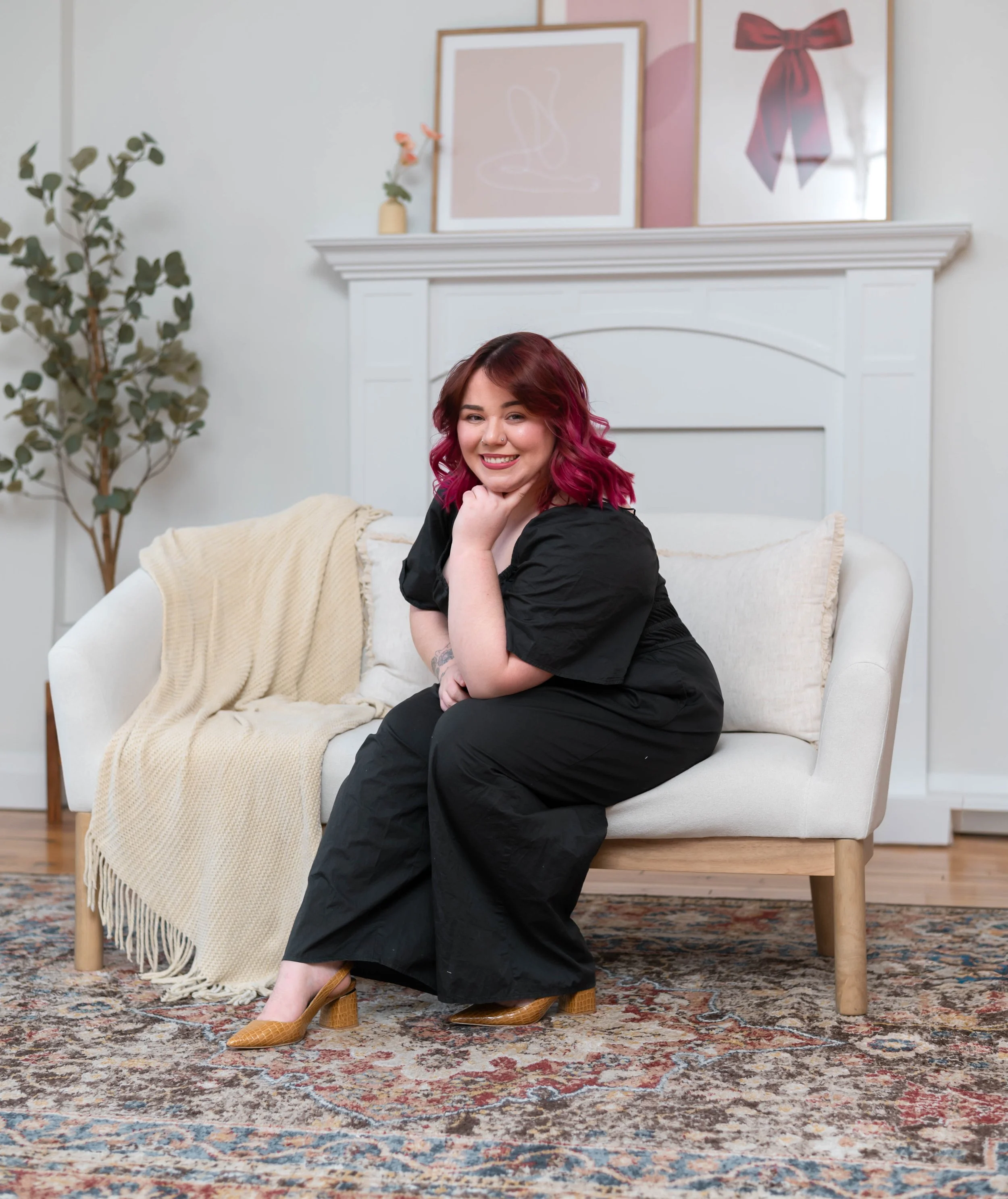 A woman with pink hair in a black dress sitting on a cream-colored sofa in a living room. Behind her, a white fireplace mantel with three framed artworks and a small vase with flowers. To the left, a potted plant. The floor has a colorful patterned rug, and a blonde blanket is draped over the sofa.