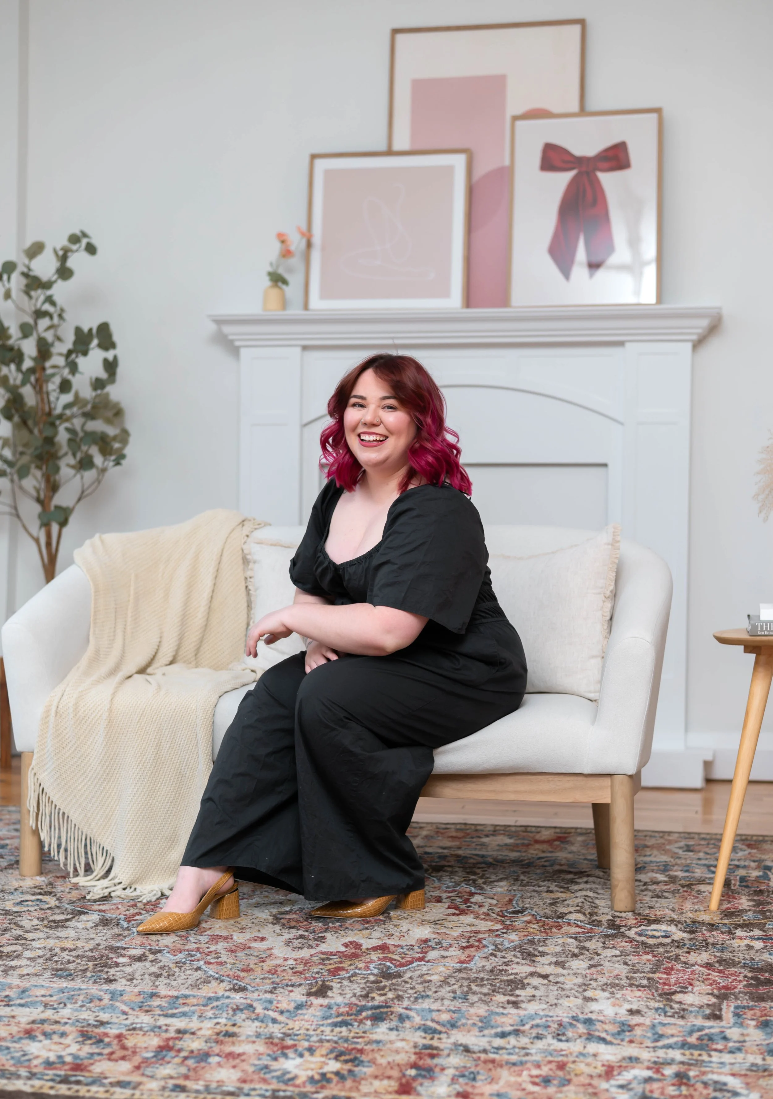 A smiling woman with pinkish-red hair sitting on a white sofa with beige blankets, wearing black clothing and brown high-heeled shoes, in a living room with framed artwork above the fireplace, a green potted plant, and a patterned rug.