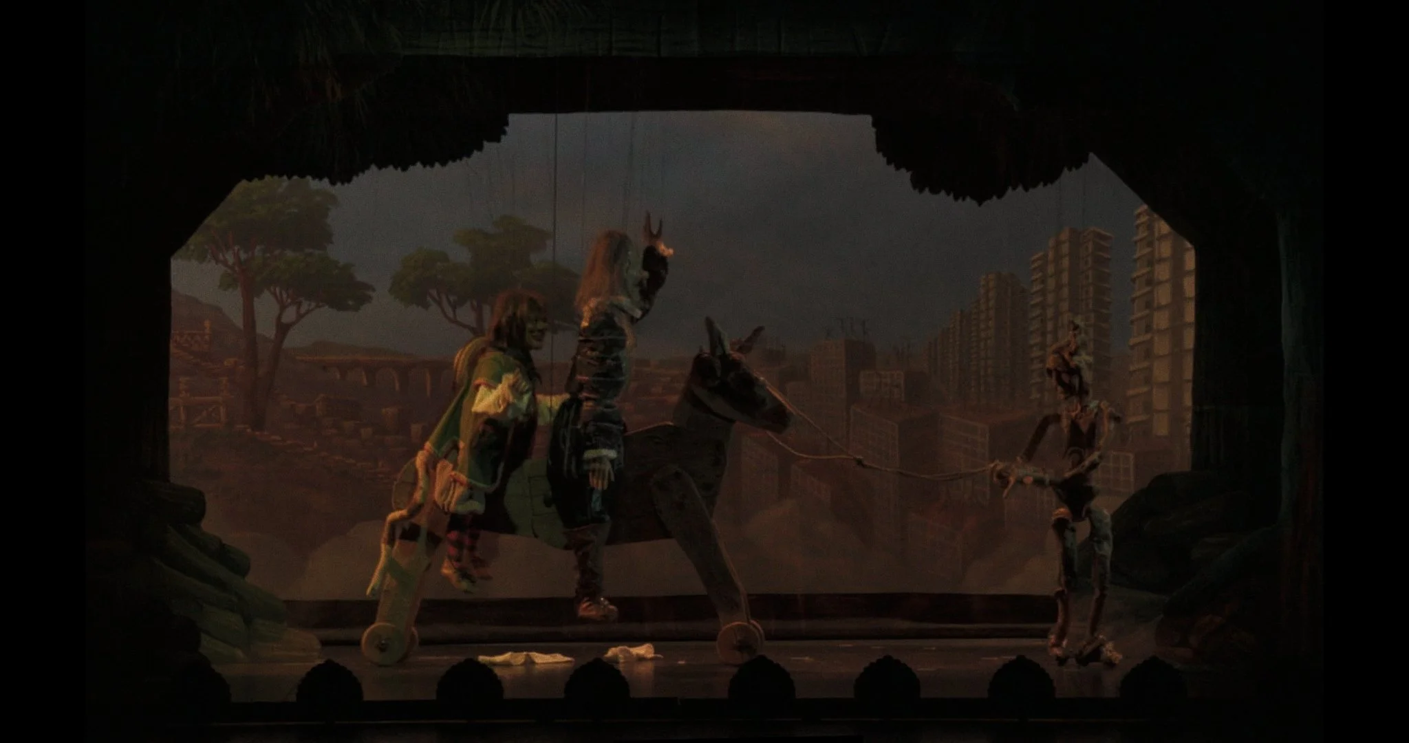 Stage marionette scene with puppets of children riding a dinosaur, a person in striped clothing, and a person on a skateboard, with cityscape and trees background.