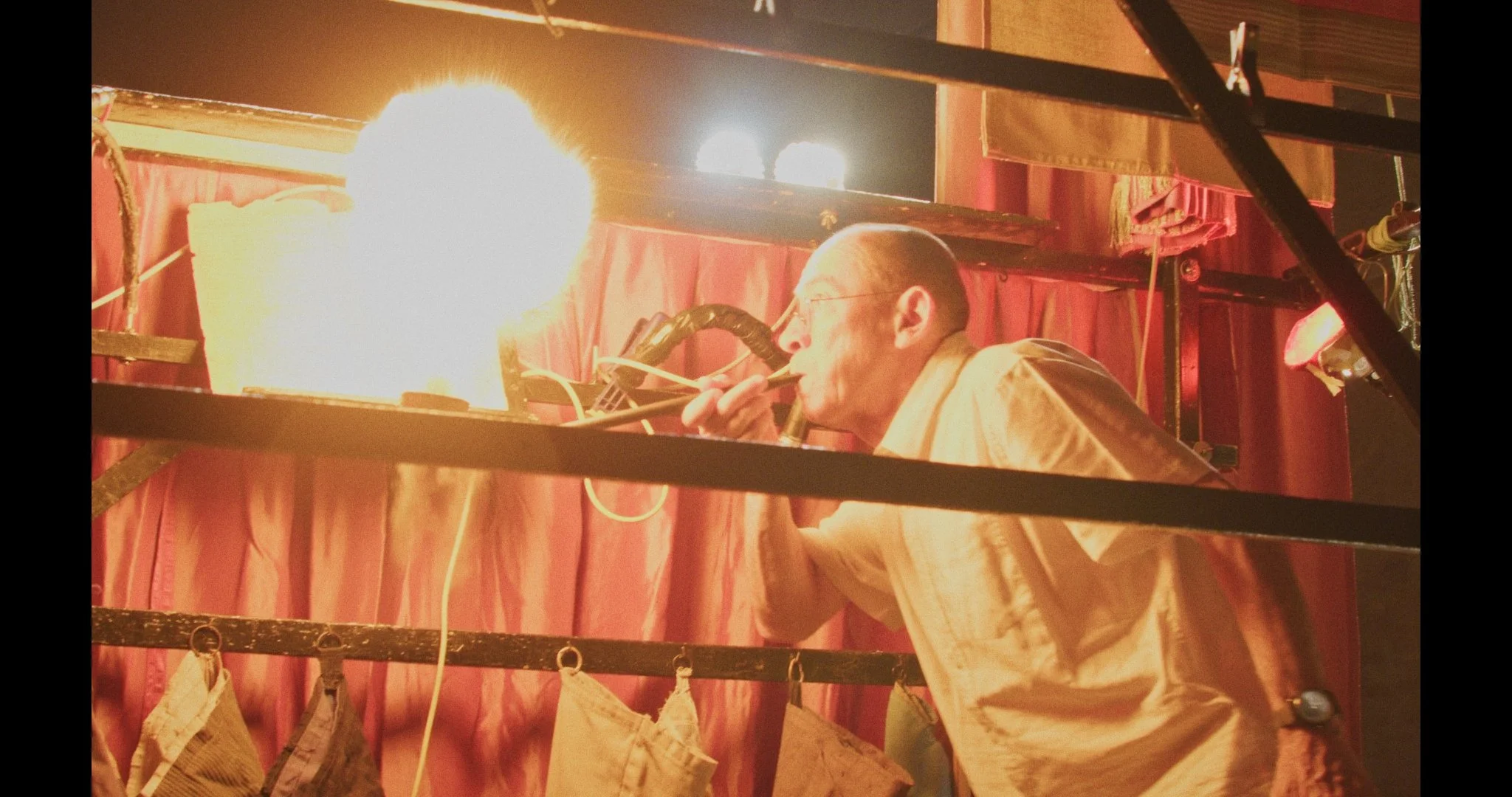 A puppeteer wearing glasses and a beige jacket creates a flame through a pipe inside a puppet theater with red curtains, wooden shelves, and hanging bags, illuminated by bright lights and a fire or explosion in the background.