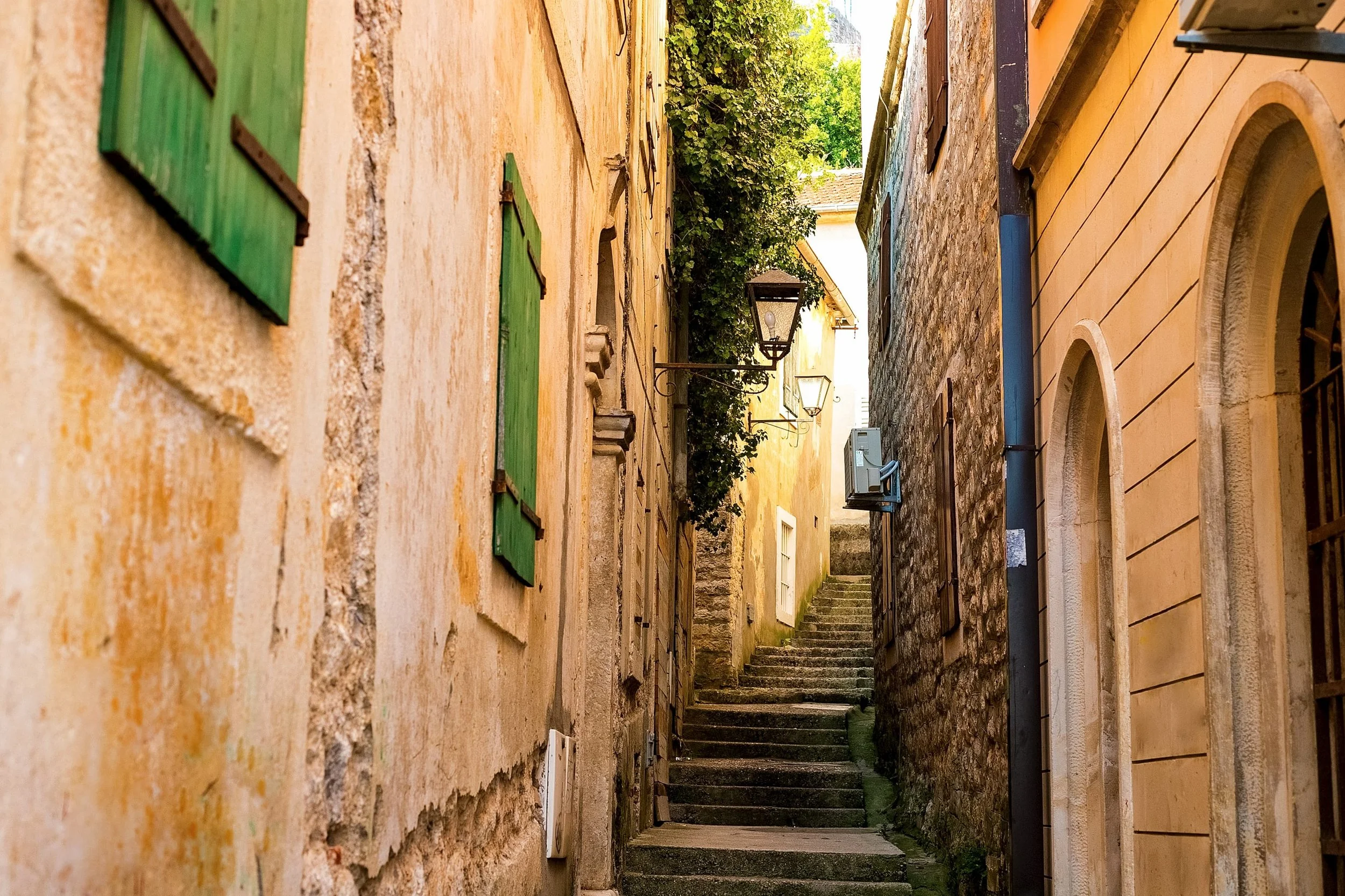 Narrow alleyway between old stone buildings with green shutters and a streetlamp, with trees visible at the end.
