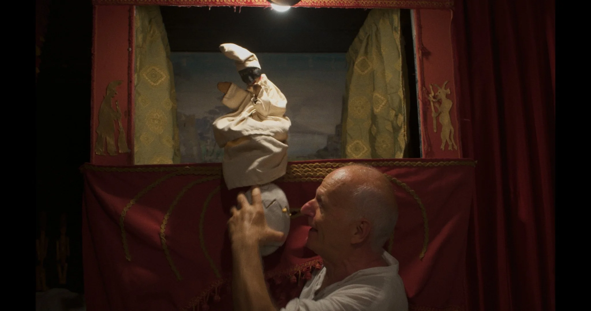 A Pulcinella puppet show where the puppeteer is interacting with the puppet in front of the stage, which is decorated with red fabric, gold trim, and gold silhouette figures, with the scenery depicted in the background.