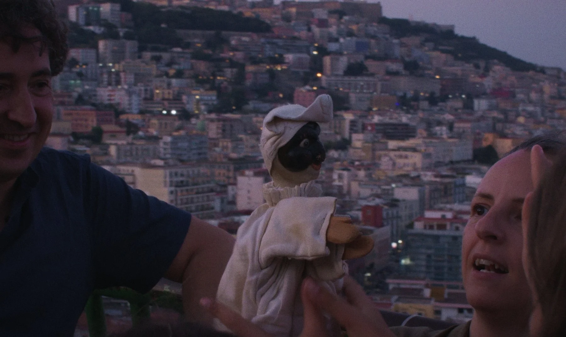 Three people and a puppet – Pulcinella – with a black face, white hat, and beige clothing outdoors in Naples at dusk.