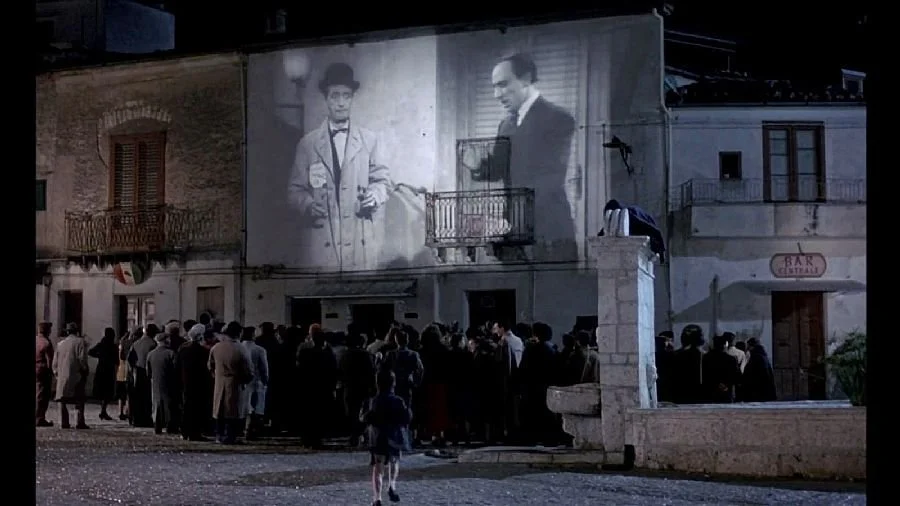 People gathered outdoors at night, watching a large black and white movie projected onto the side of a building.