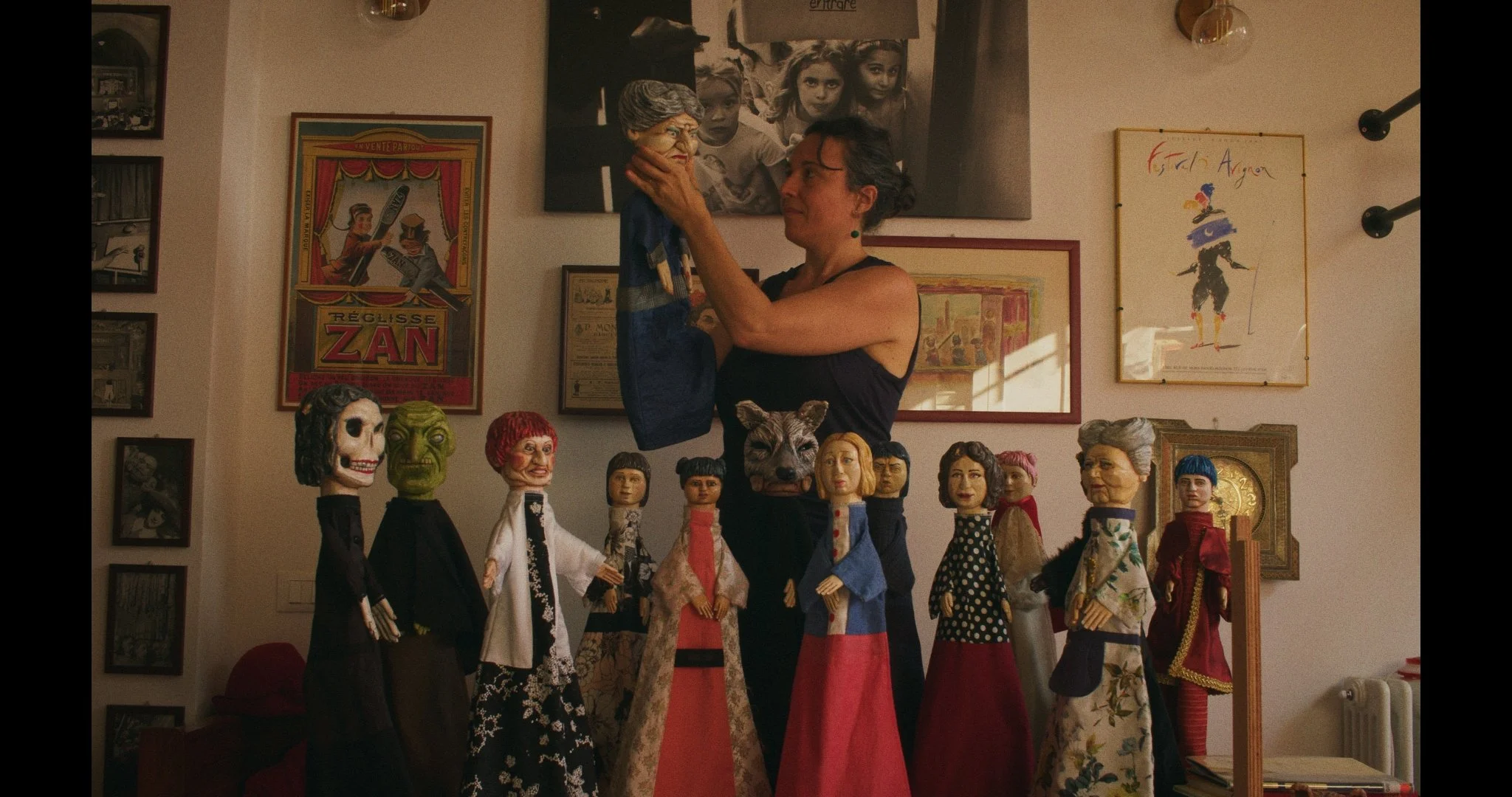 A puppeteer a puppet that looks like an elderly woman with gray hair and glasses, surrounded by various handcrafted puppets and dolls on a table. The background features framed posters and photographs on the wall.