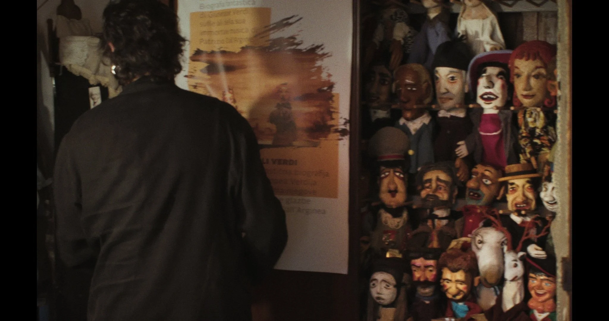 A puppeteer with dark curly hair wearing a black jacket, facing a display of puppets, with a colorful poster on the wall behind them.