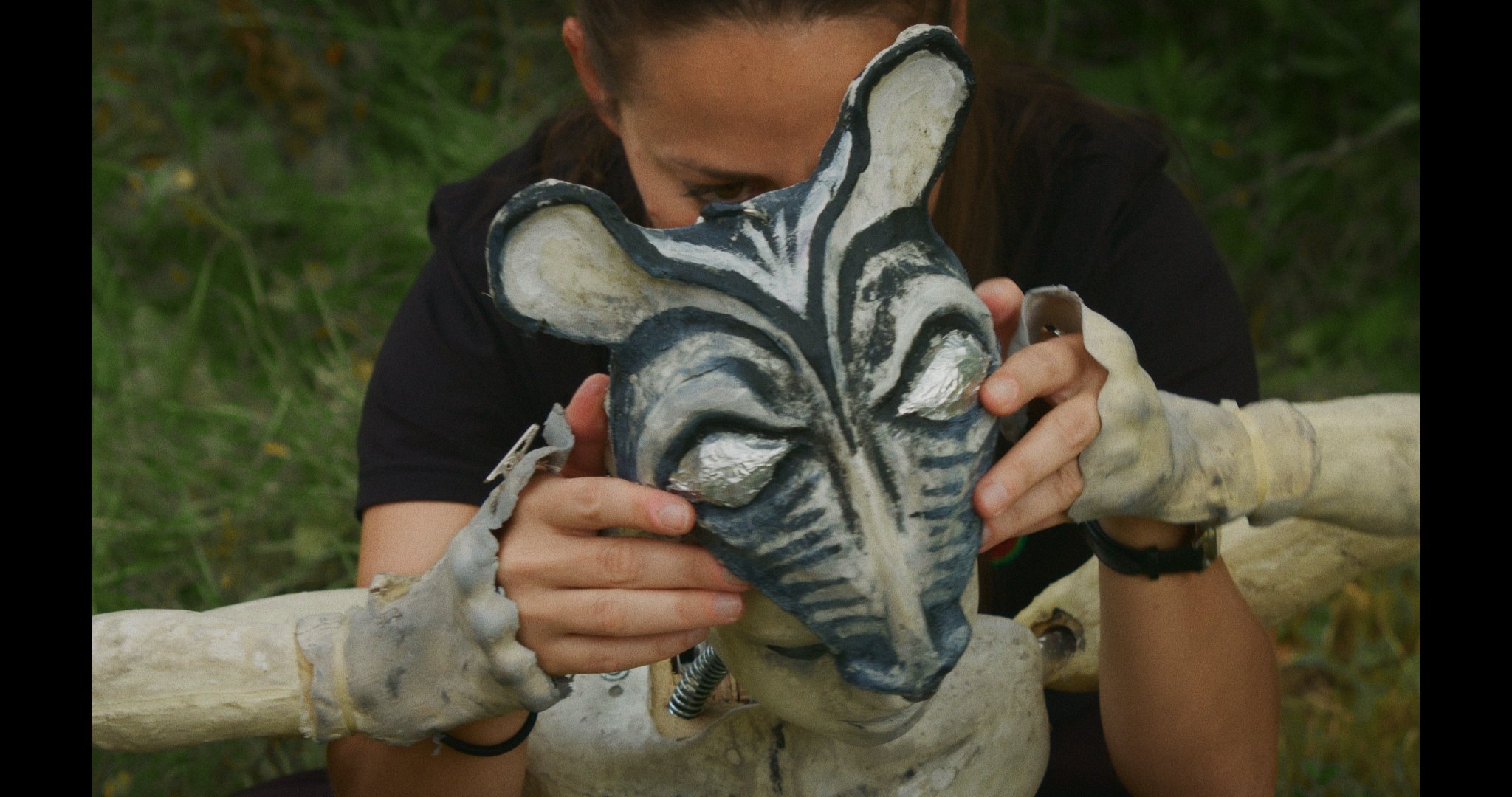A puppeteer holding a detailed zebra-shaped mask, partially covering their face, outdoors with green foliage in the background.