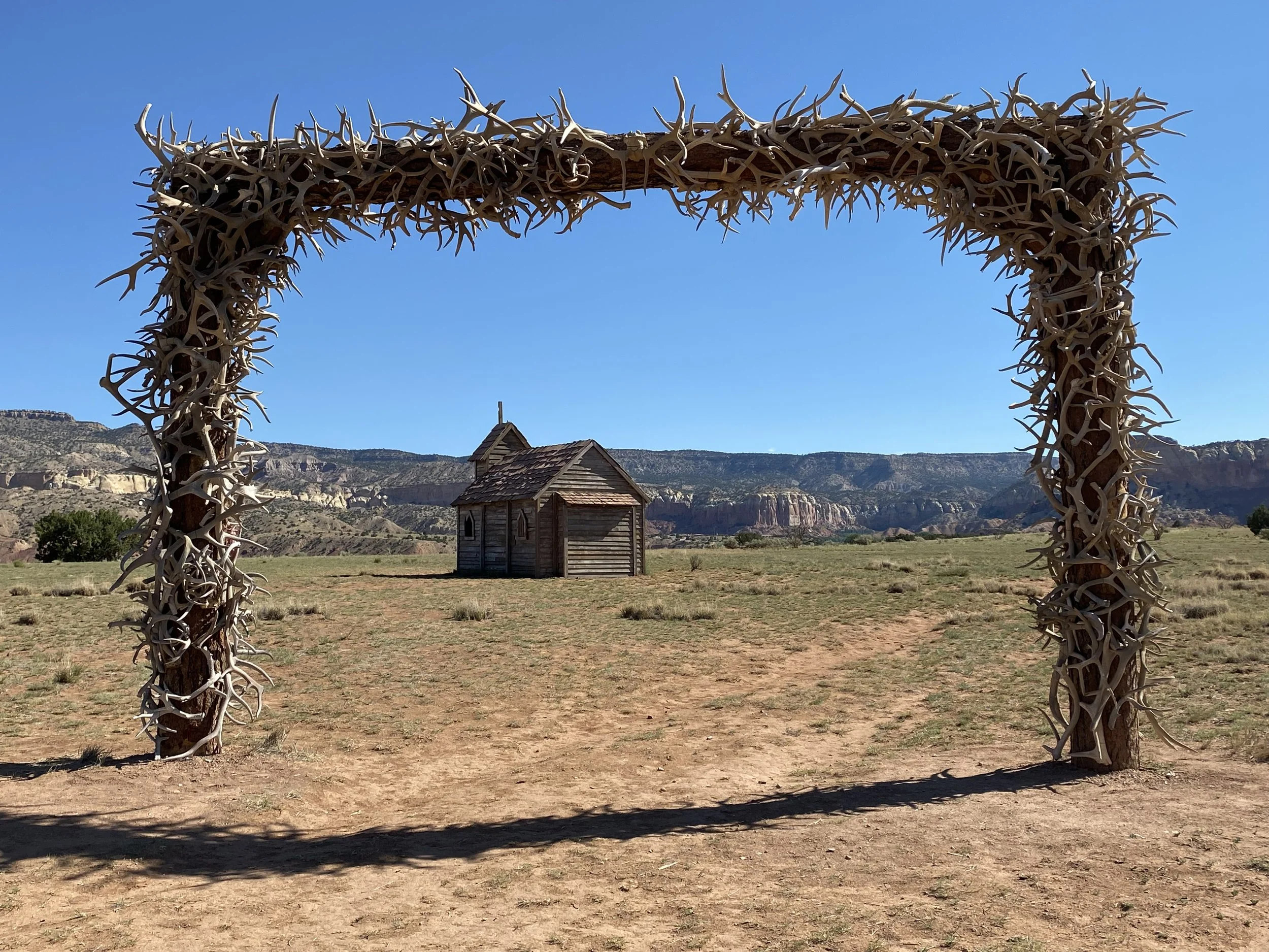 A wooden arch covered with antlers frames a small wooden house in a desert landscape with distant rocky cliffs and clear blue sky.