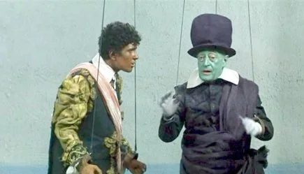 Two actors presenting to be marionettes: one is a young man, Othello, in colorful clothing and the other is an older man, Jago, a performer costume with a painted green face, top hat, and dark outfit.