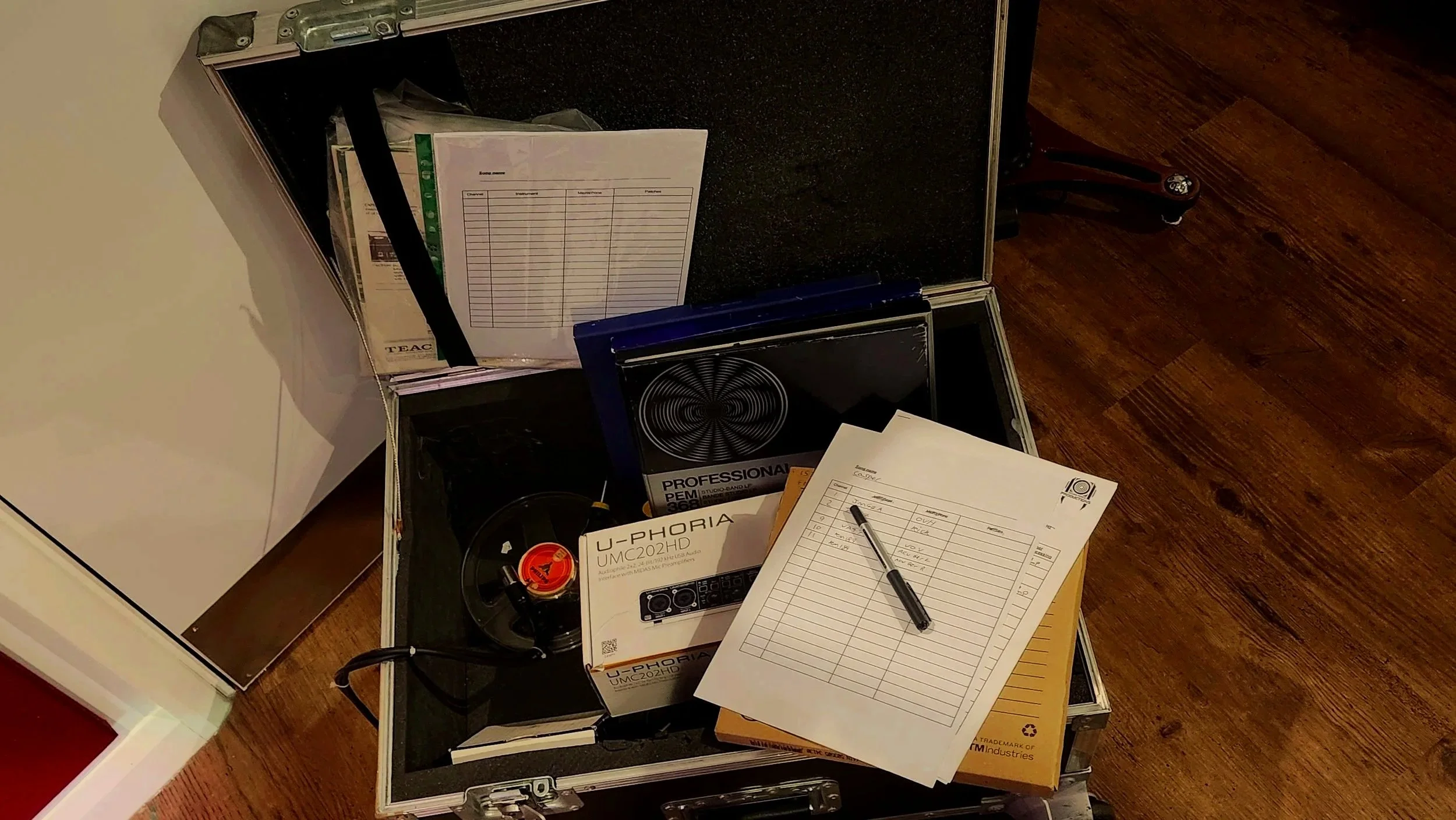Open black equipment case filled with music and work-related items, including a music speaker, documents, and a pen, on hardwood floor.