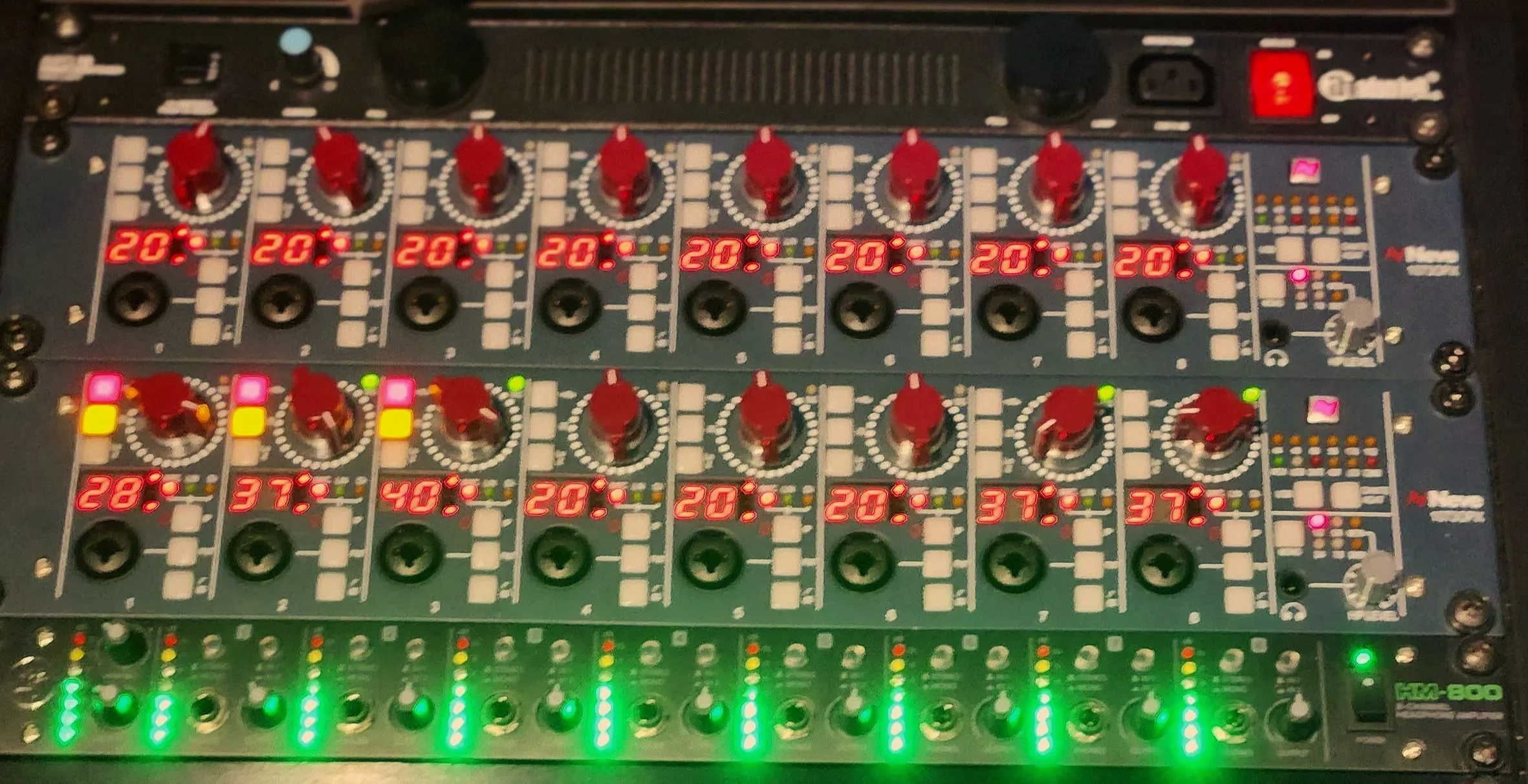 An audio mixing console with multiple knobs, sliders, and digital displays showing temperature or level readings, along with various colored indicator lights.