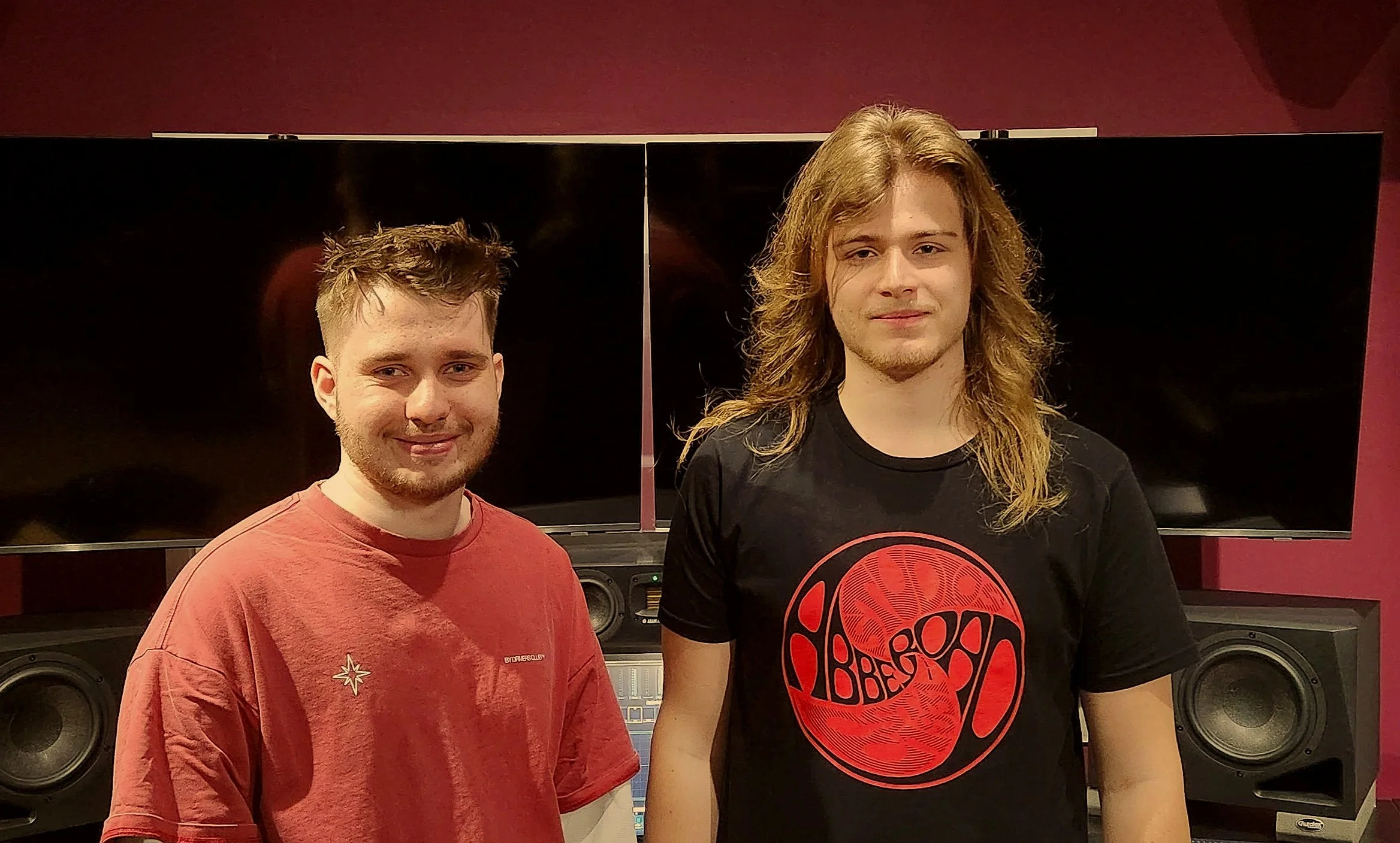 Two young men standing in a music studio, one wearing a red shirt and the other a black shirt with a red circular design, in front of audio speakers and mixing equipment.