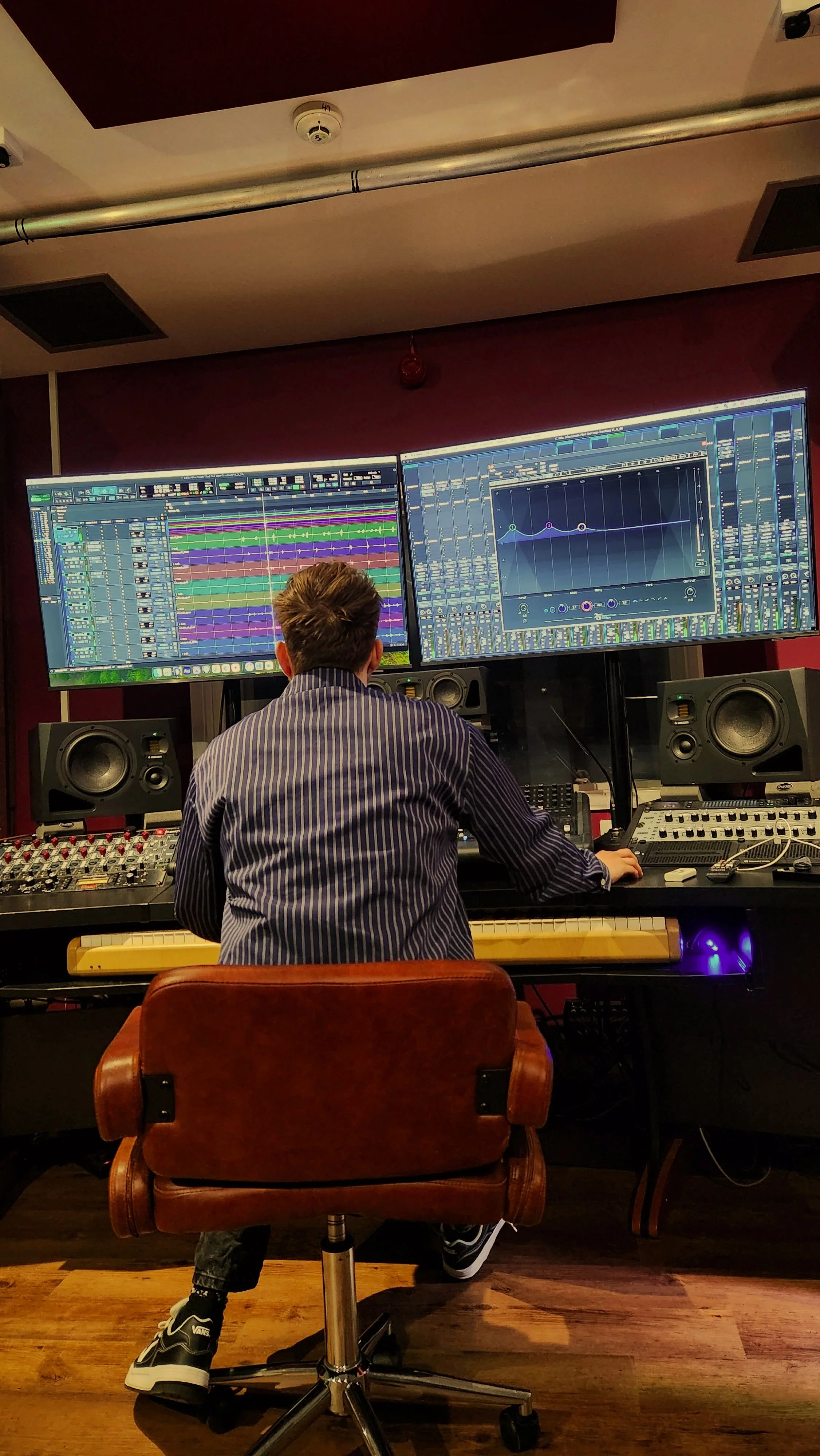 A person with short hair, wearing a striped shirt and sneakers, is working in a music studio at a large mixing console with two big computer monitors displaying digital audio workstations, surrounded by speakers and audio equipment.