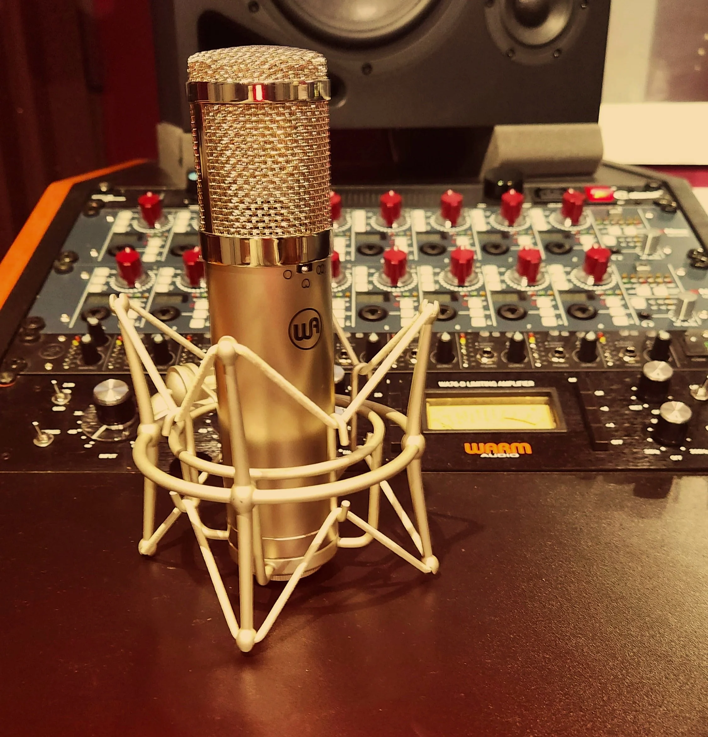 A gold-colored microphone with a metal mesh grille is mounted on a table with a mixer and audio equipment in the background.