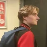 Side profile of a young man with medium-length blond hair, wearing a red jacket and carrying a backpack, standing in front of a dark door or elevator.