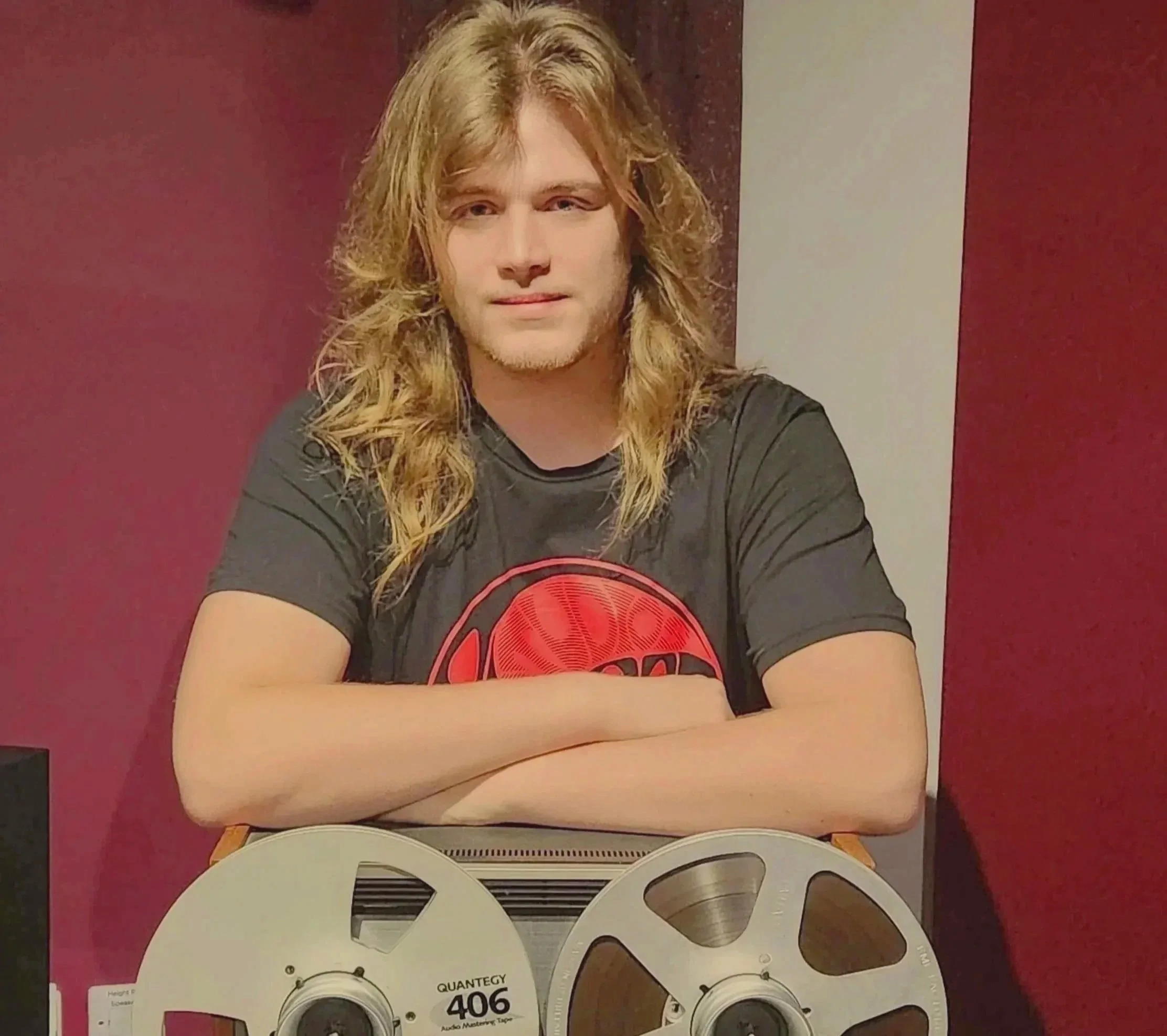 A young man with long, wavy blond hair and a light beard, wearing a black T-shirt with a red graphic design, sits at a table with his arms crossed, in front of a maroon and white background.
