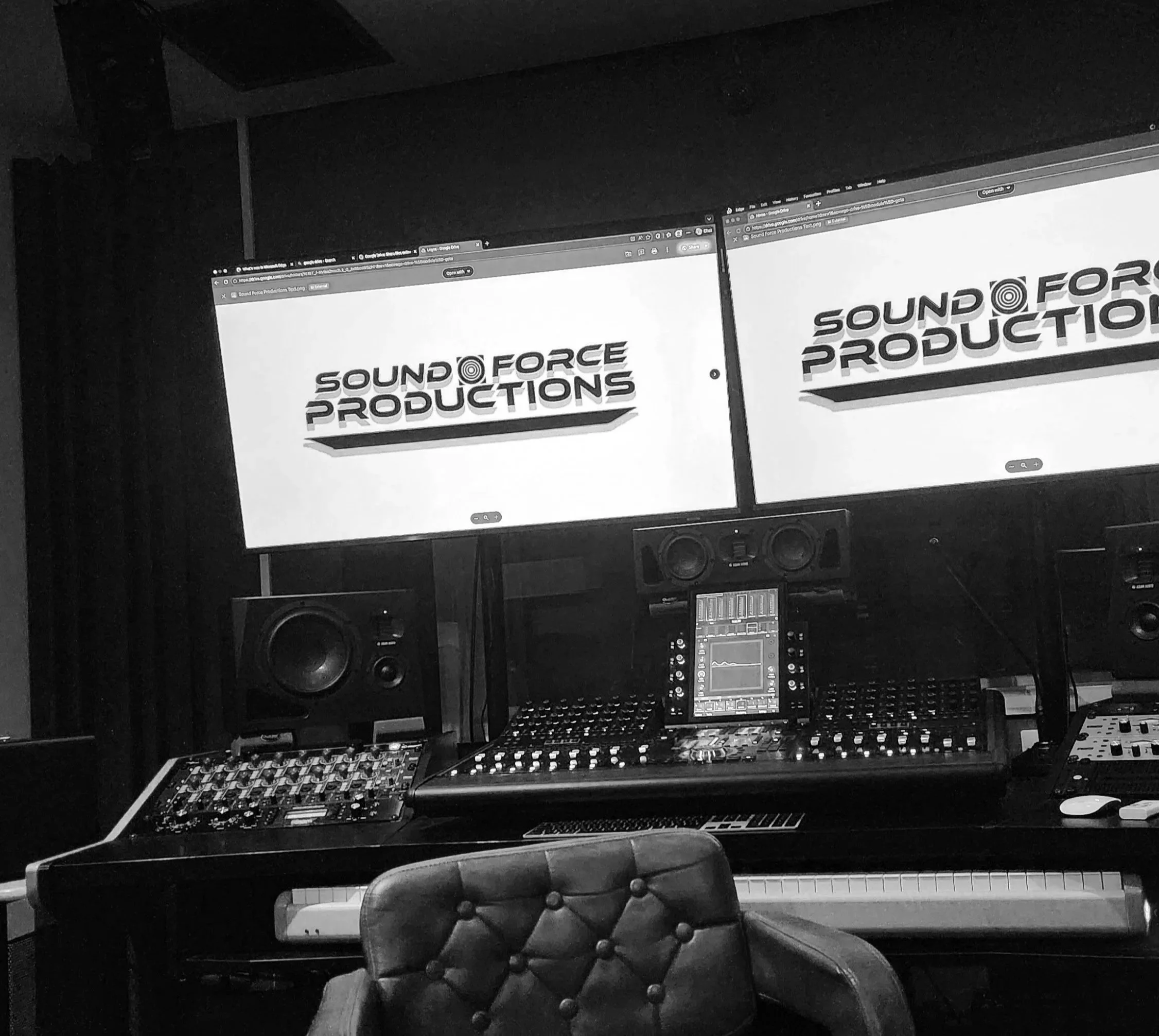 Photo of a music studio workspace with two computer monitors displaying the Sound Force Productions logo, surrounded by audio equipment, speakers, keyboard, and a chair.