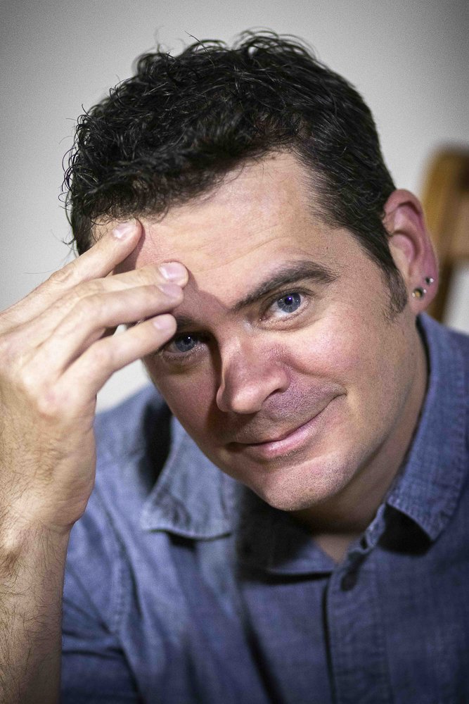 A man with dark curly hair and blue eyes, wearing a denim shirt, smiling and holding his hand to his forehead in a thinking pose.