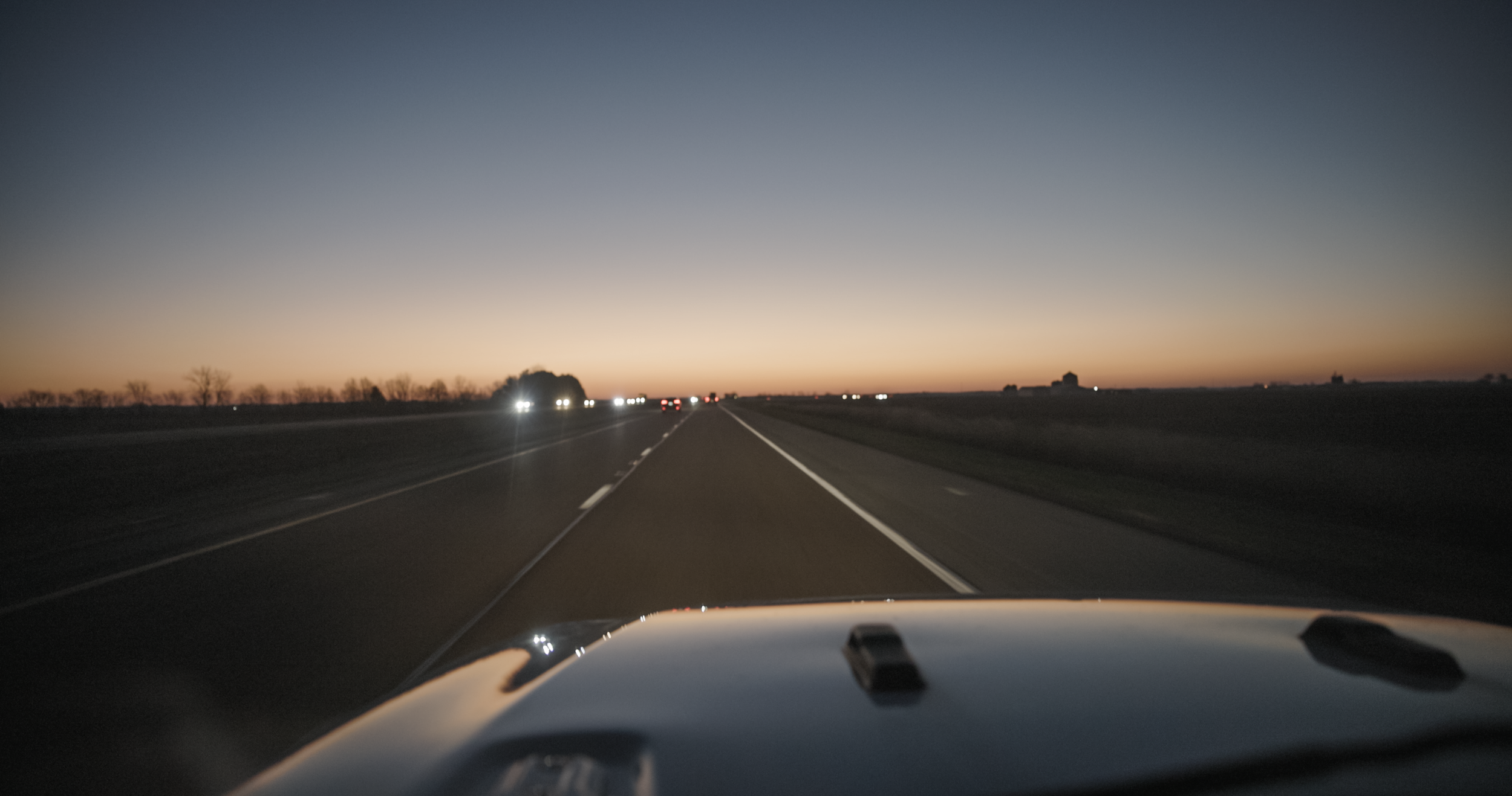 View of a highway at dusk with a car front visible, surrounded by open fields and dimly lit houses in the distance.
