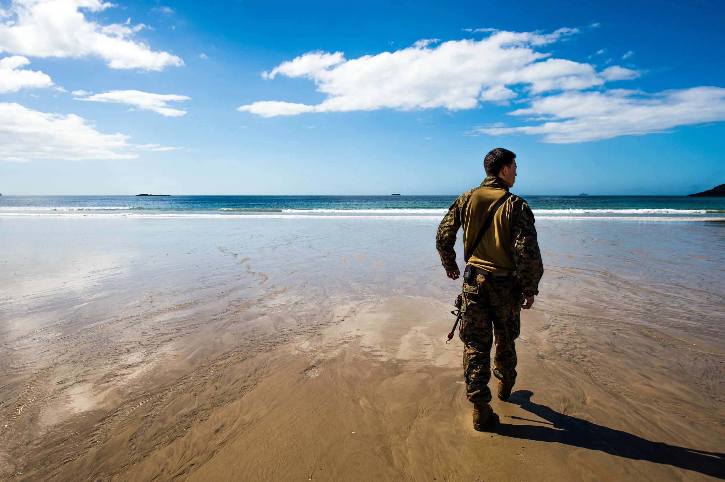 A man in camouflage military uniform walking on a sandy beach near the ocean under a partly cloudy sky.