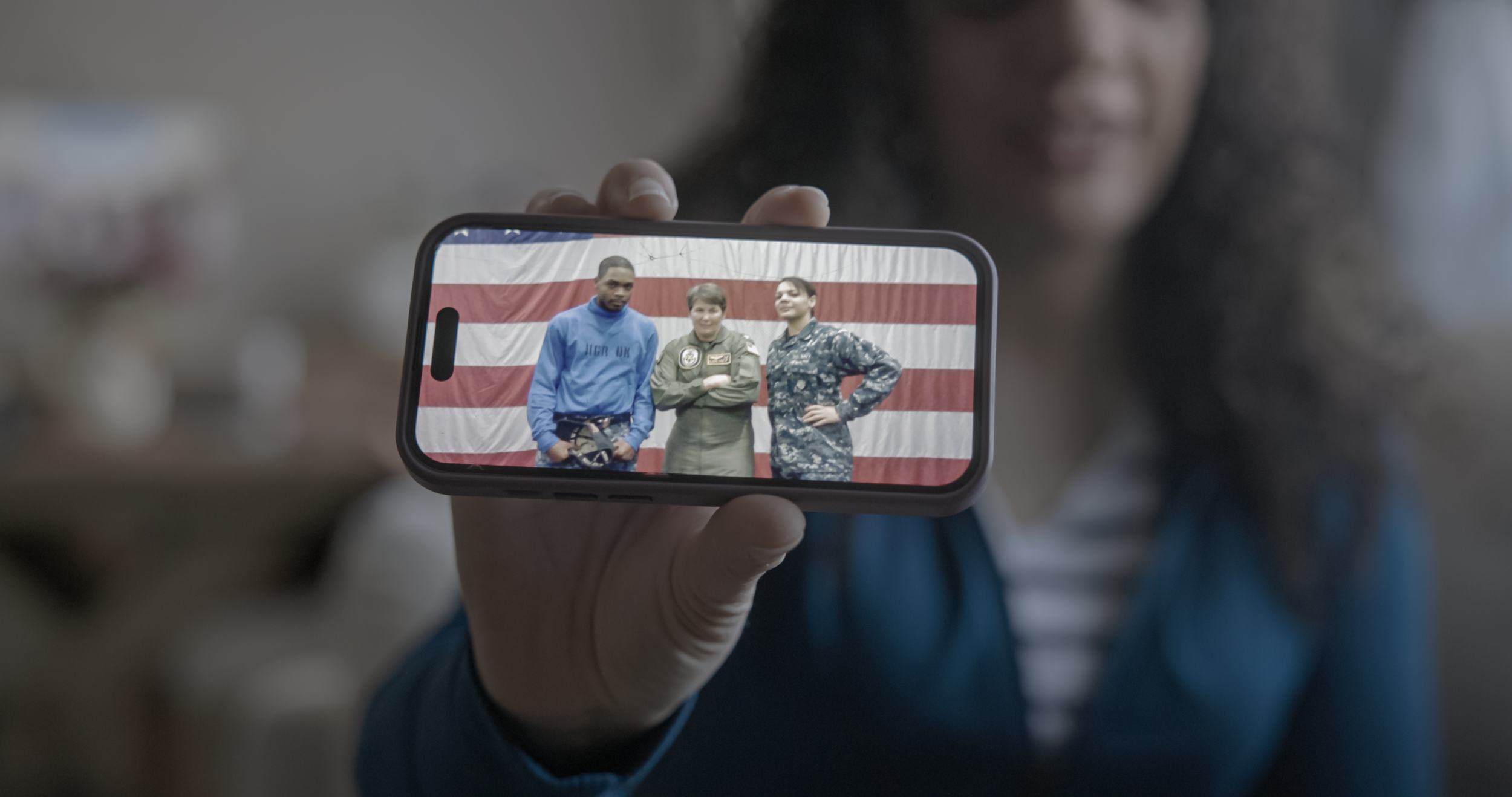 Person holding a smartphone displaying three individuals standing in front of an American flag.