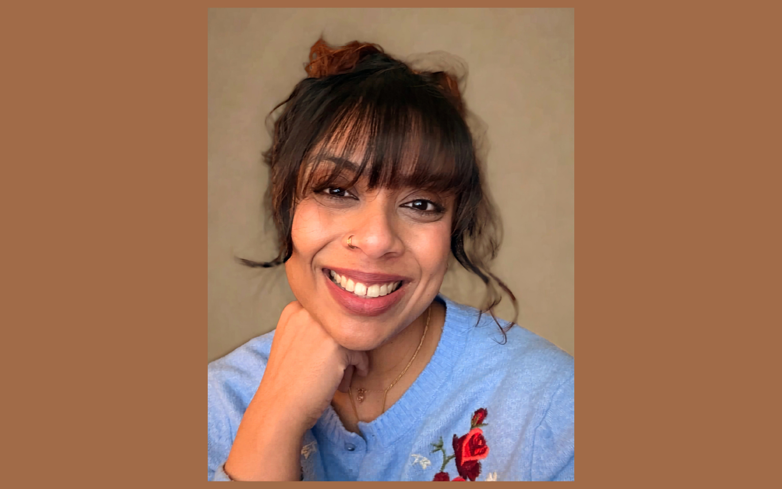 A woman with dark hair styled in a messy bun, wearing a light blue top with floral embroidery, and smiling with her teeth showing. She has a nose ring and is resting her chin on her hand.