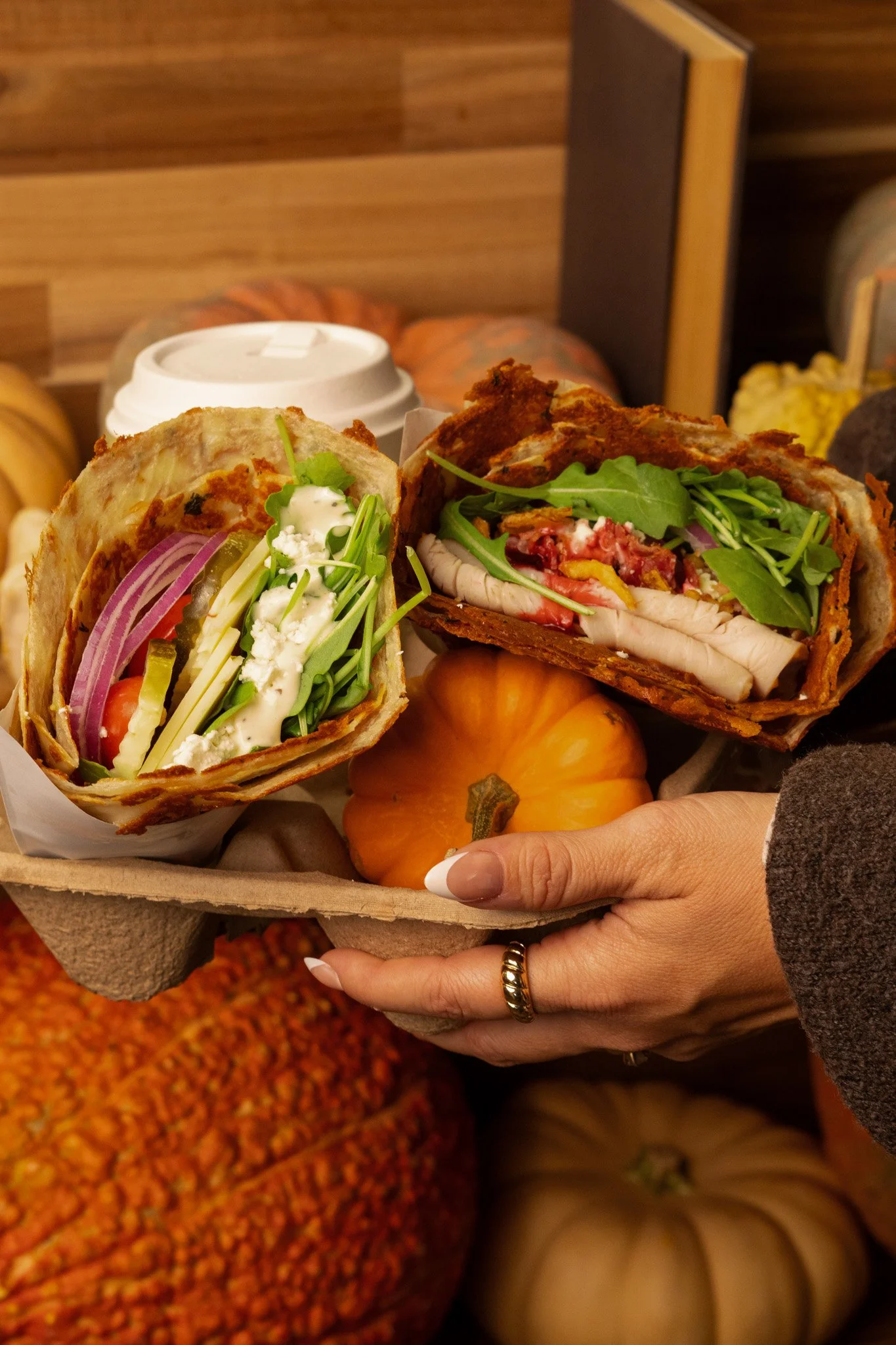 A person holding a tray with two sandwiches filled with lettuce, tomato, red onion, cheese, and meat, surrounded by small pumpkins.