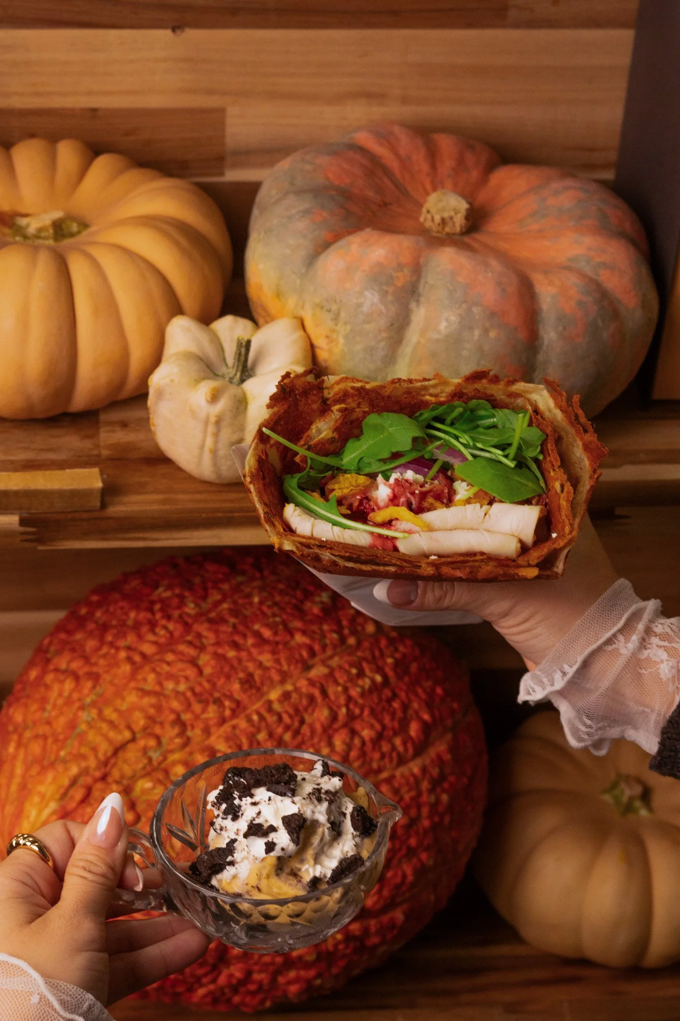A person holding a glass bowl of ice cream with Oreo cookies, whipped cream, and a sandwich wrap, with a background of pumpkins and a person's lap in colorful, textured clothing.