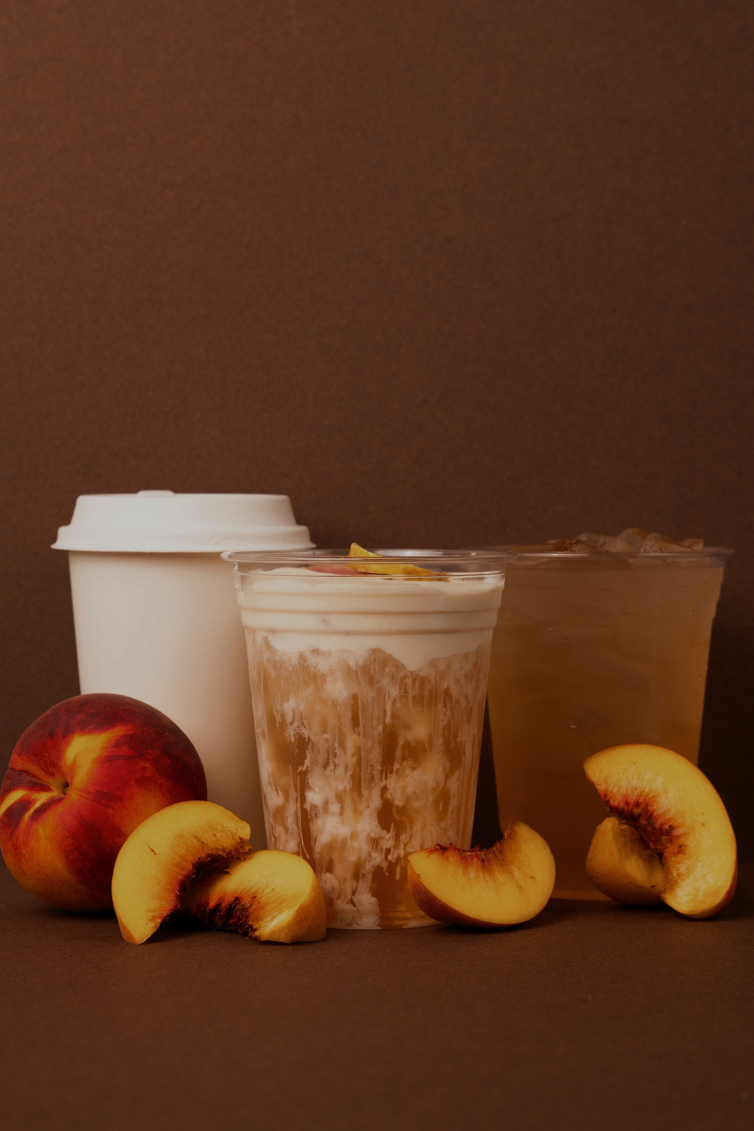A plastic cup with a iced yerba mate drink, a white to-go coffee cup, and several peaches, some sliced. The background is brown.
