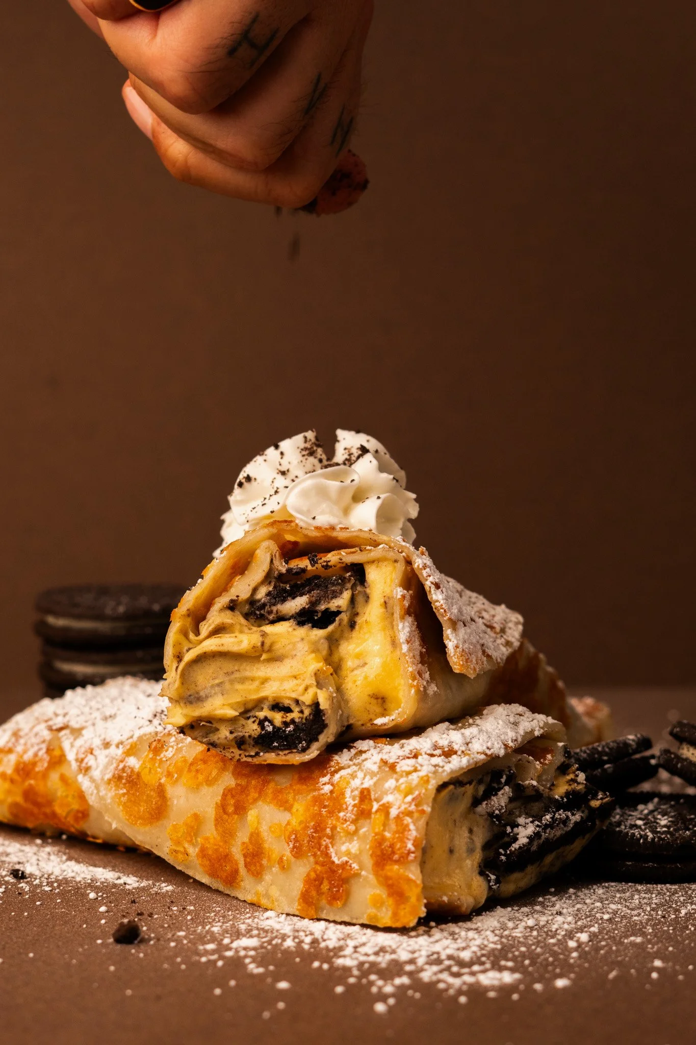 A slice of cookies and cream cheesecake desert roll with whipped cream topping and chocolate crumbs, dusted with powdered sugar, on a dark surface with another dessert in the background.