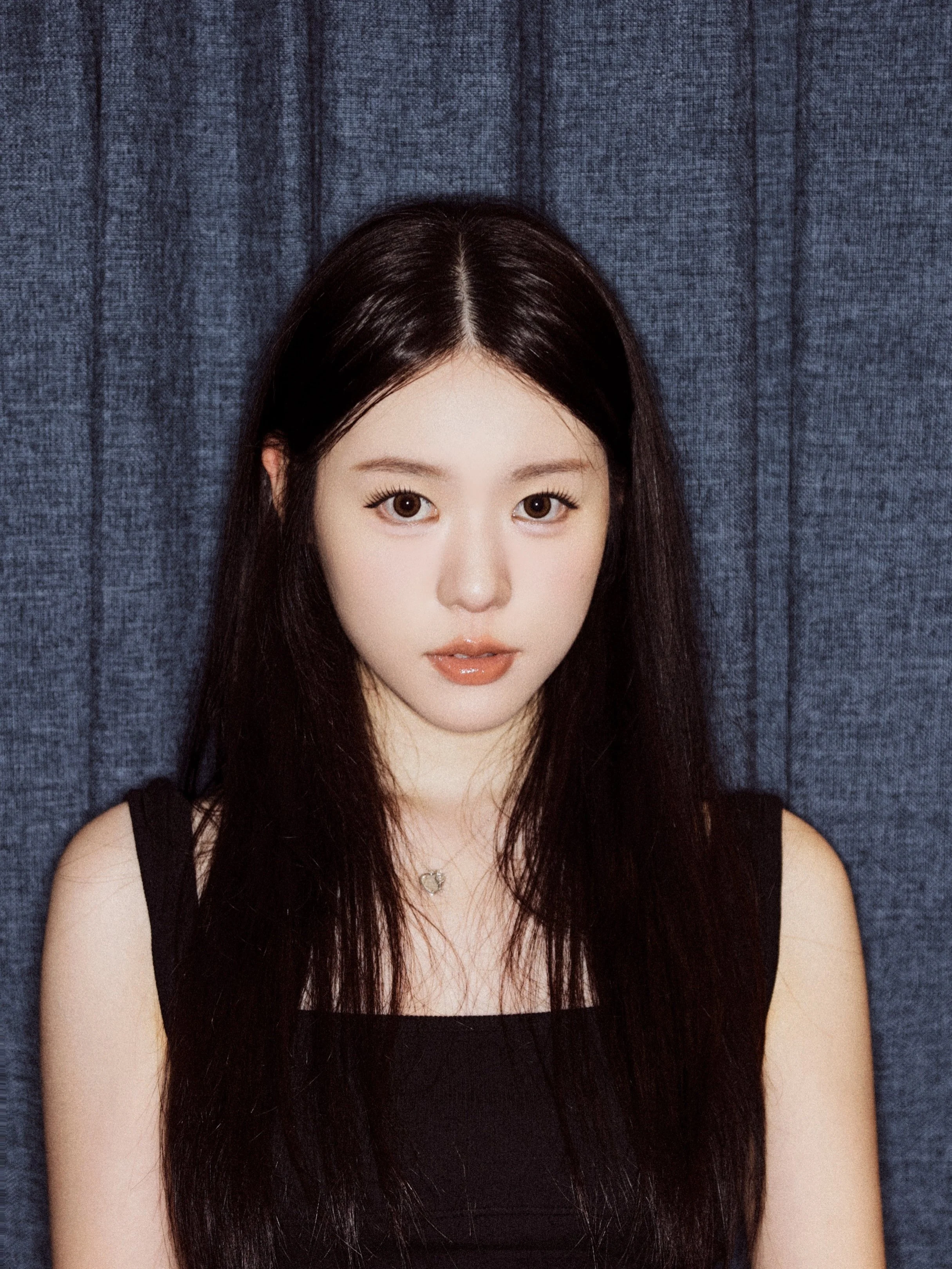 A woman with long dark hair, wearing a black sleeveless top, standing in front of a dark blue curtain.