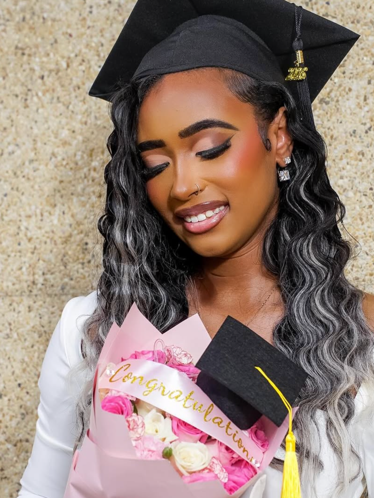 A woman in a graduation cap holding a bouquet of pink and white flowers with a 'Congratulations' ribbon, smiling with eyes closed.
