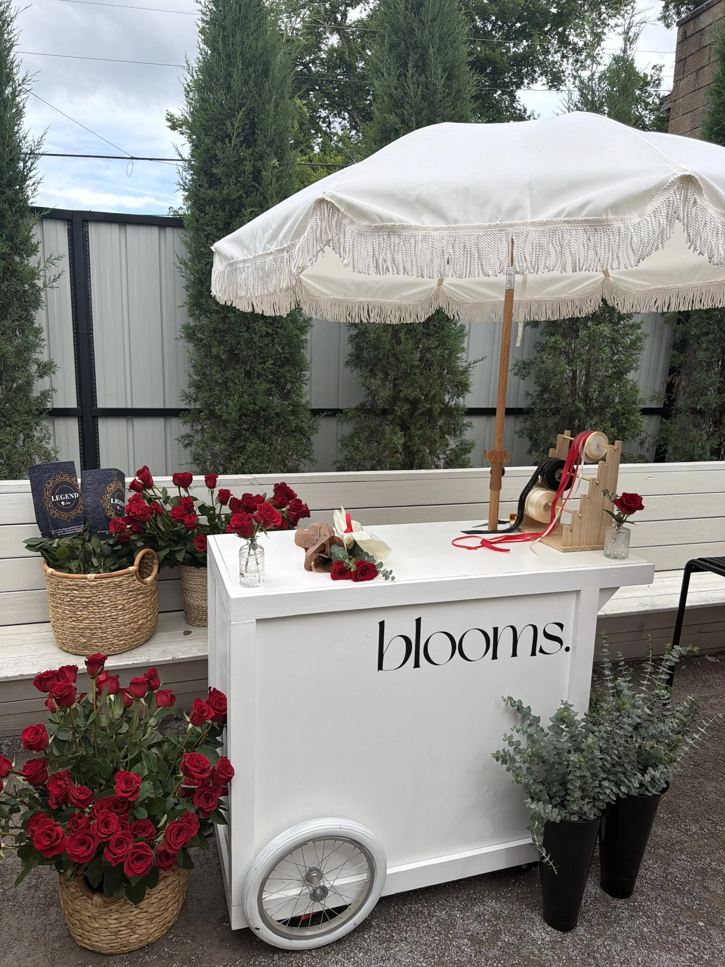 A flower cart with red roses, a white sign with the word 'blooms.' on it, and an umbrella. The cart is positioned outdoors against a backdrop of tall green trees and a gray metal fence. The cart has a wheel and contains decorative items and rose bouq