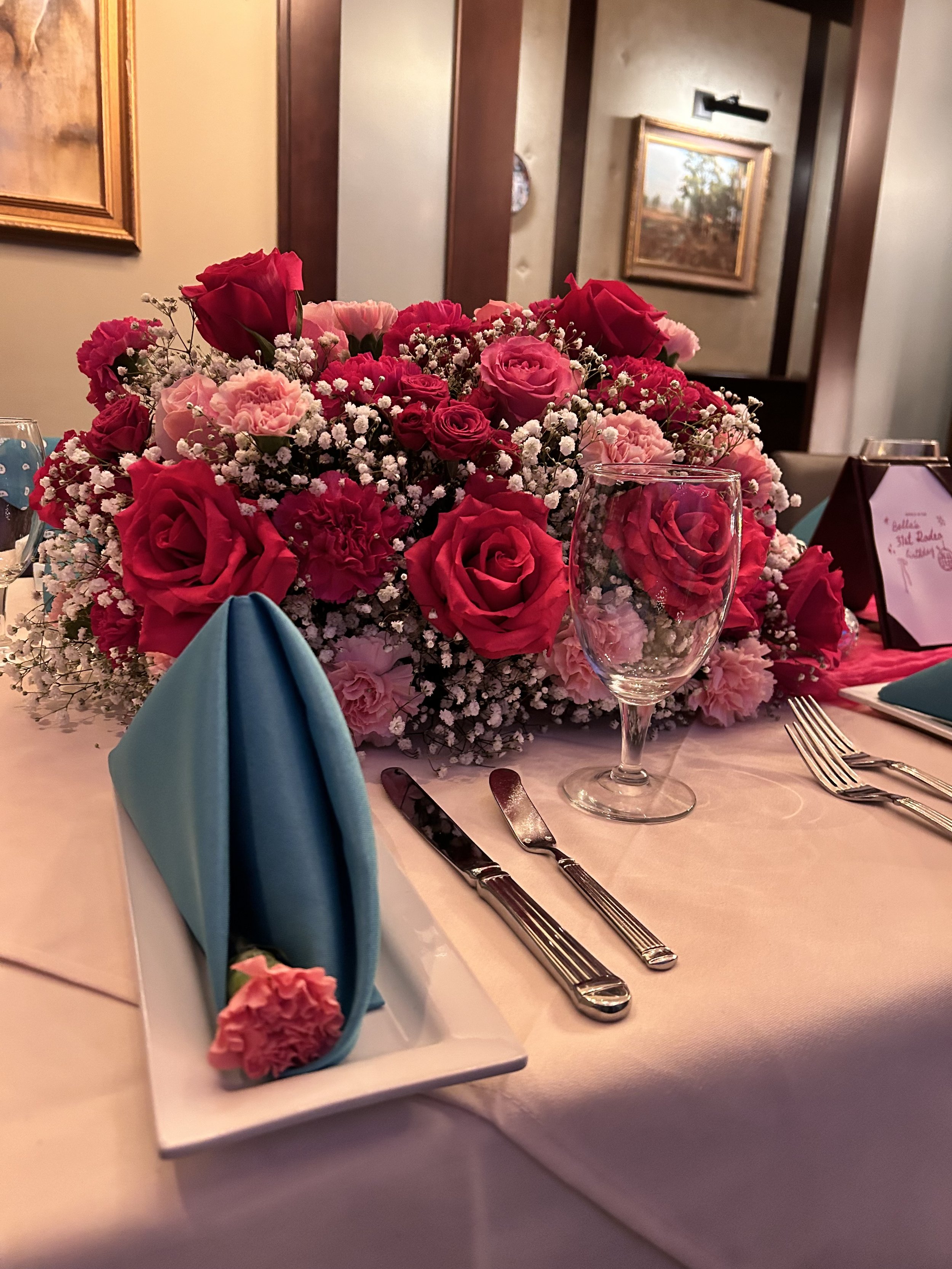 A centerpieces of pink and red roses and baby's breath on a banquet table, with blue cloth napkins, silverware, a wine glass, and a framed name card in the background.