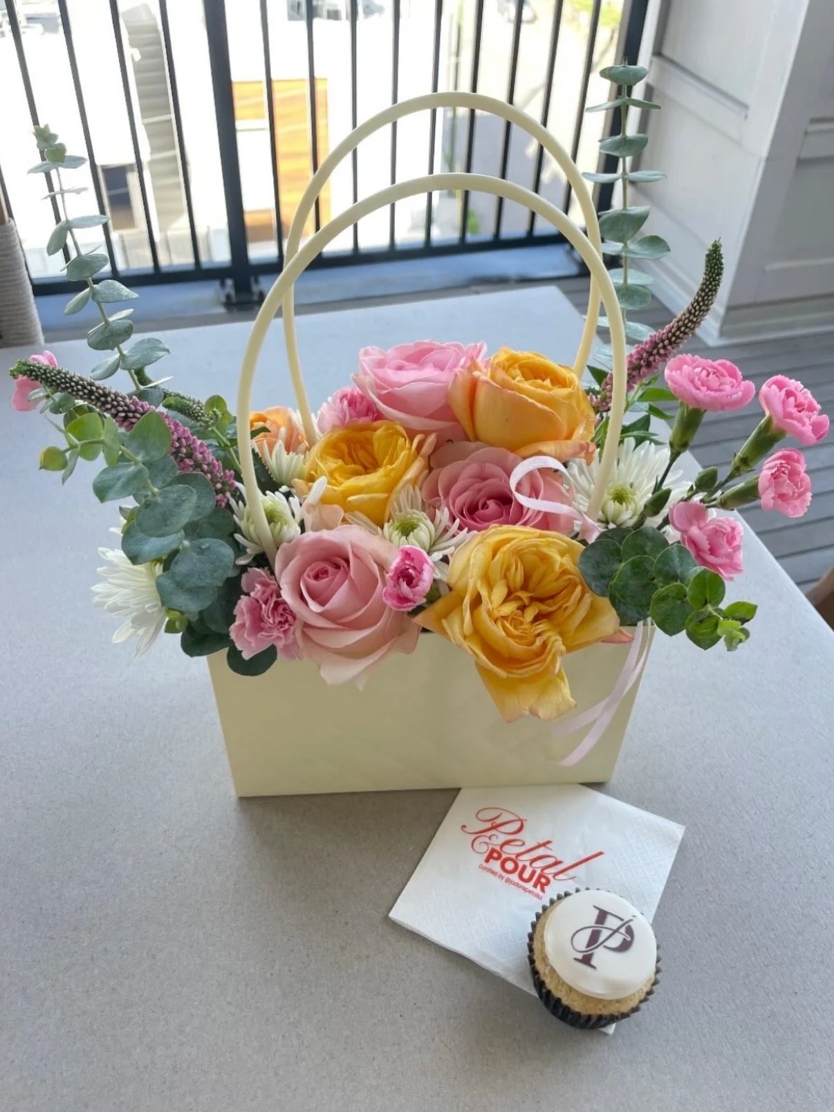 A white basket filled with pink, yellow, and peach roses, along with white daisies and eucalyptus leaves, sits on a table with a cupcake and a 