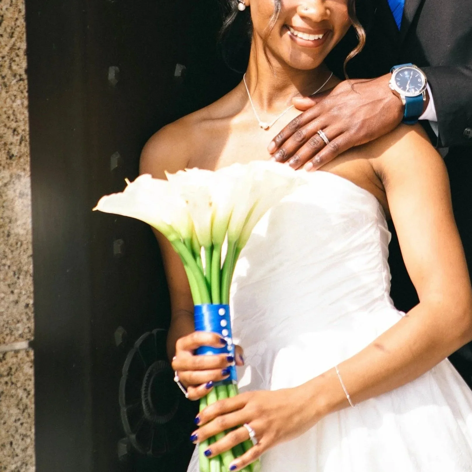 A woman in a strapless white dress smiling while holding a bouquet of white calla lilies. A man's hand with a ring and a watch rests on her shoulder.