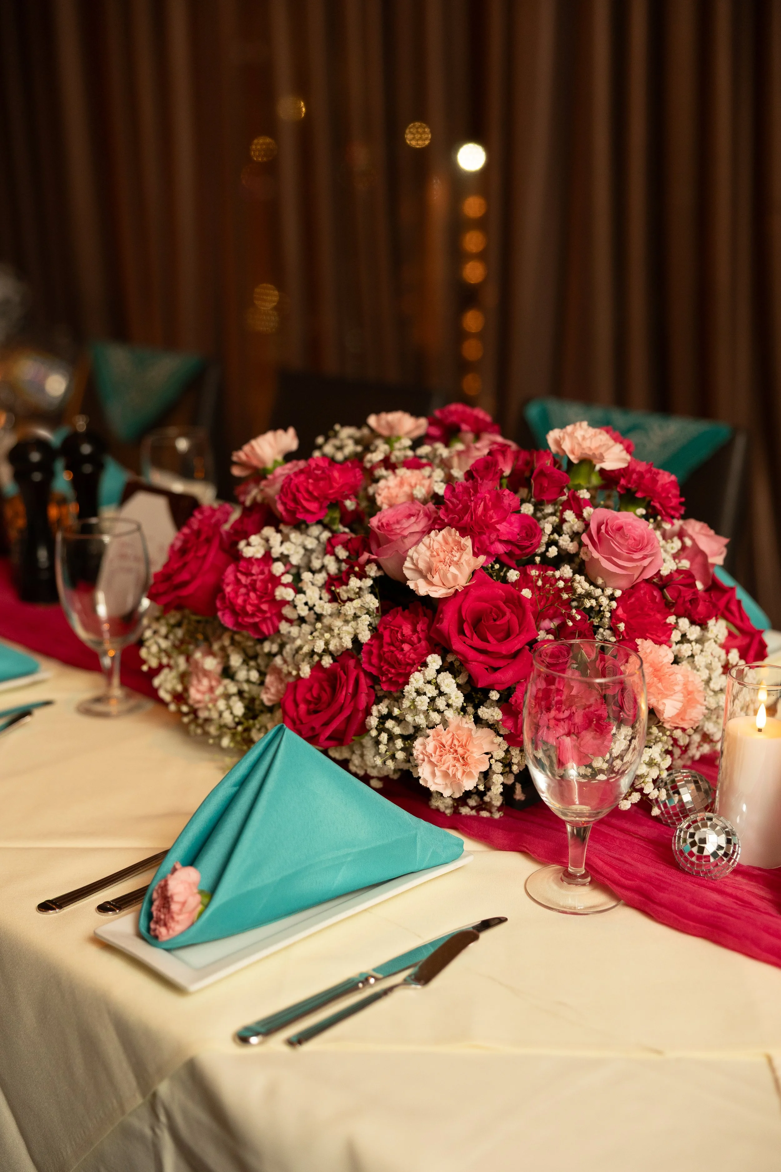 A table decorated for a formal event with a large floral centerpiece of pink and white flowers, turquoise napkins on white plates, crystal glasses, silverware, and a red table runner, set against a wooden background with string lights.