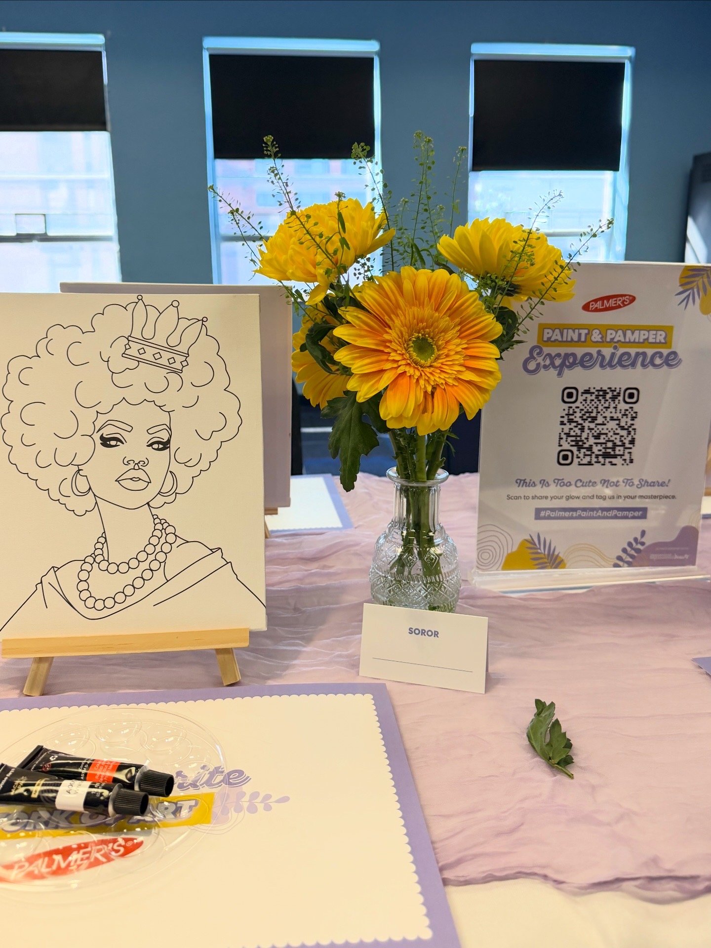 A table with a drawing of a woman with curly hair wearing a crown and pearl necklace, a vase with yellow and orange flowers, a sign with the name Soror, and promotional materials for Palmer's paint and pamper experience.