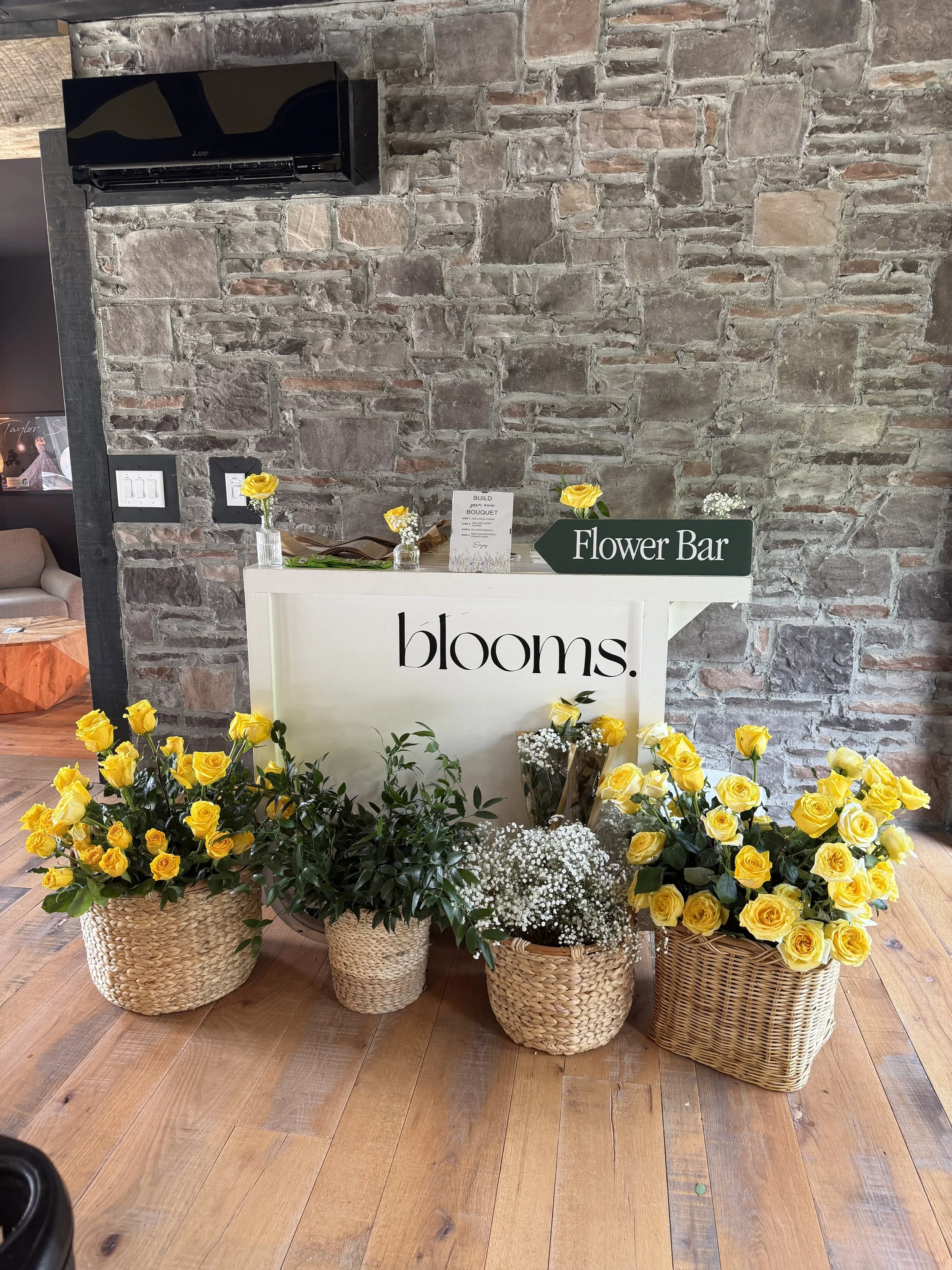 A floral display with yellow roses and small white flowers arranged in woven baskets in front of a brick wall. There is a white counter with a sign that reads 'Flower Bar' and some small vases with flowers on top.