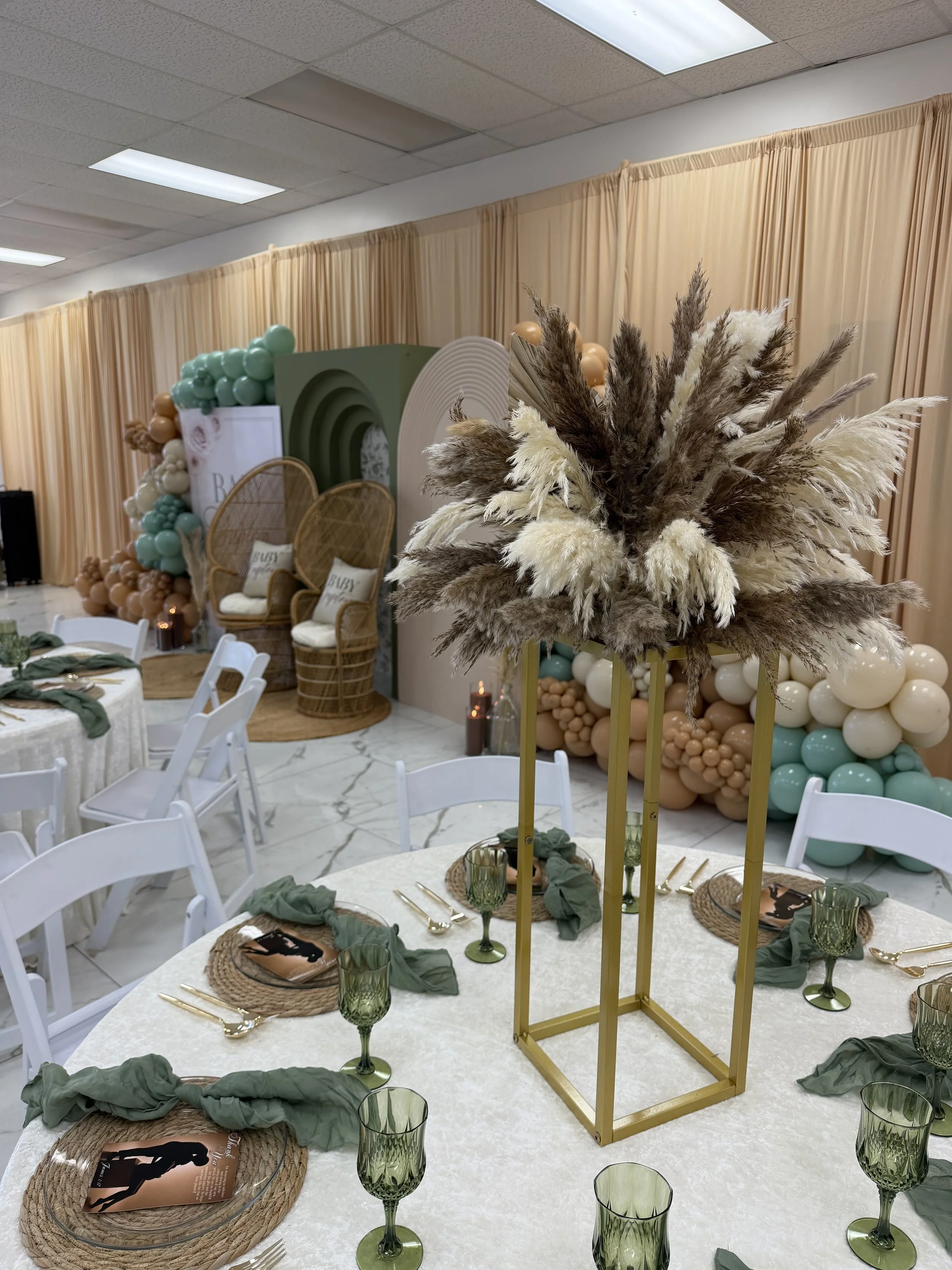 Decorated event space with a large floral arrangement of pampas grass in the foreground, round tables with olive green glasses, natural woven placemats and green napkins, a backdrop with balloons, rattan chairs, and beige curtains.