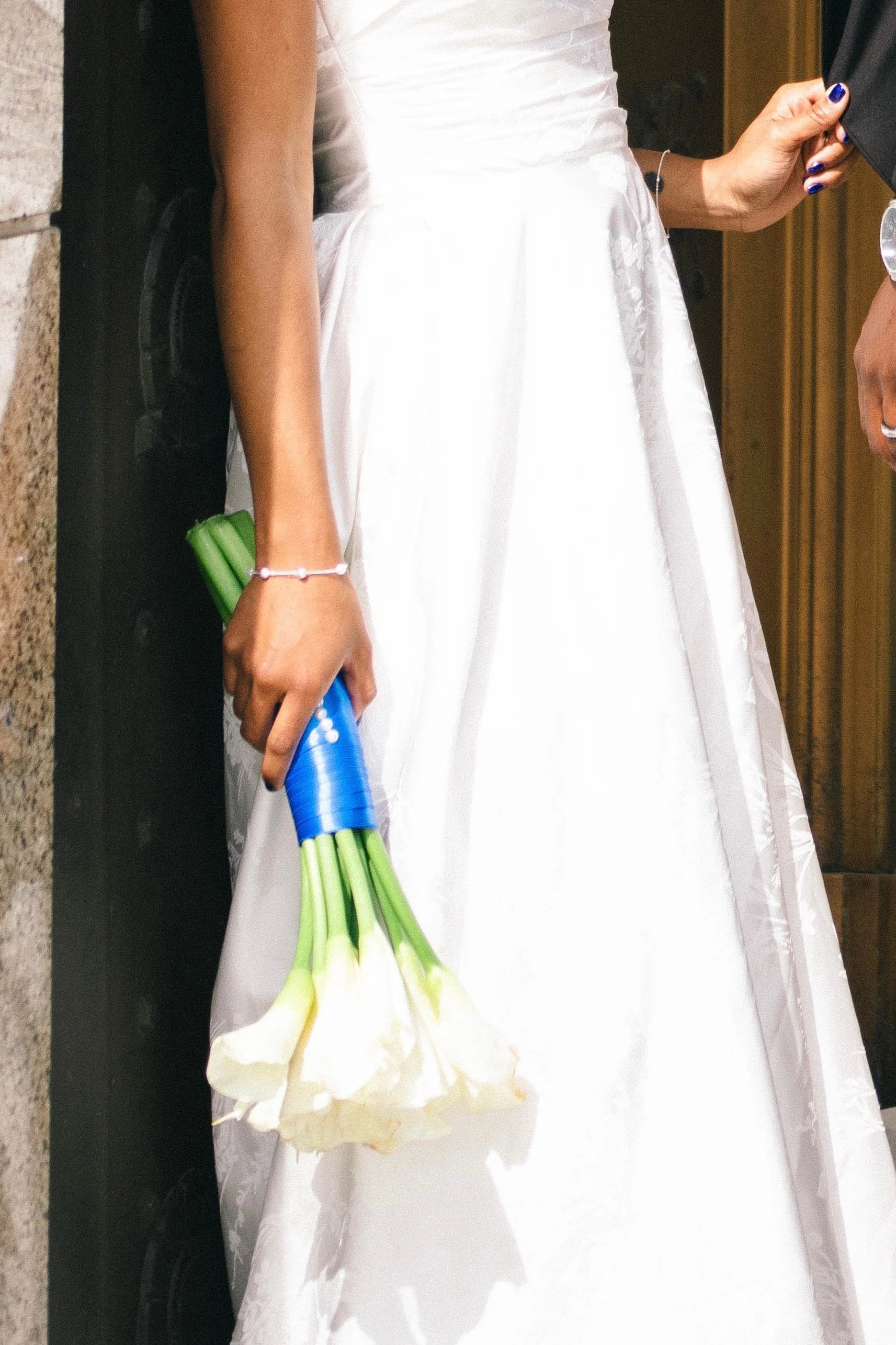 Close-up of a bride wearing a white wedding dress holding a bouquet of white calla lilies, with a hand showing blue nail polish and bracelets, standing by a black surface against a wooden background.