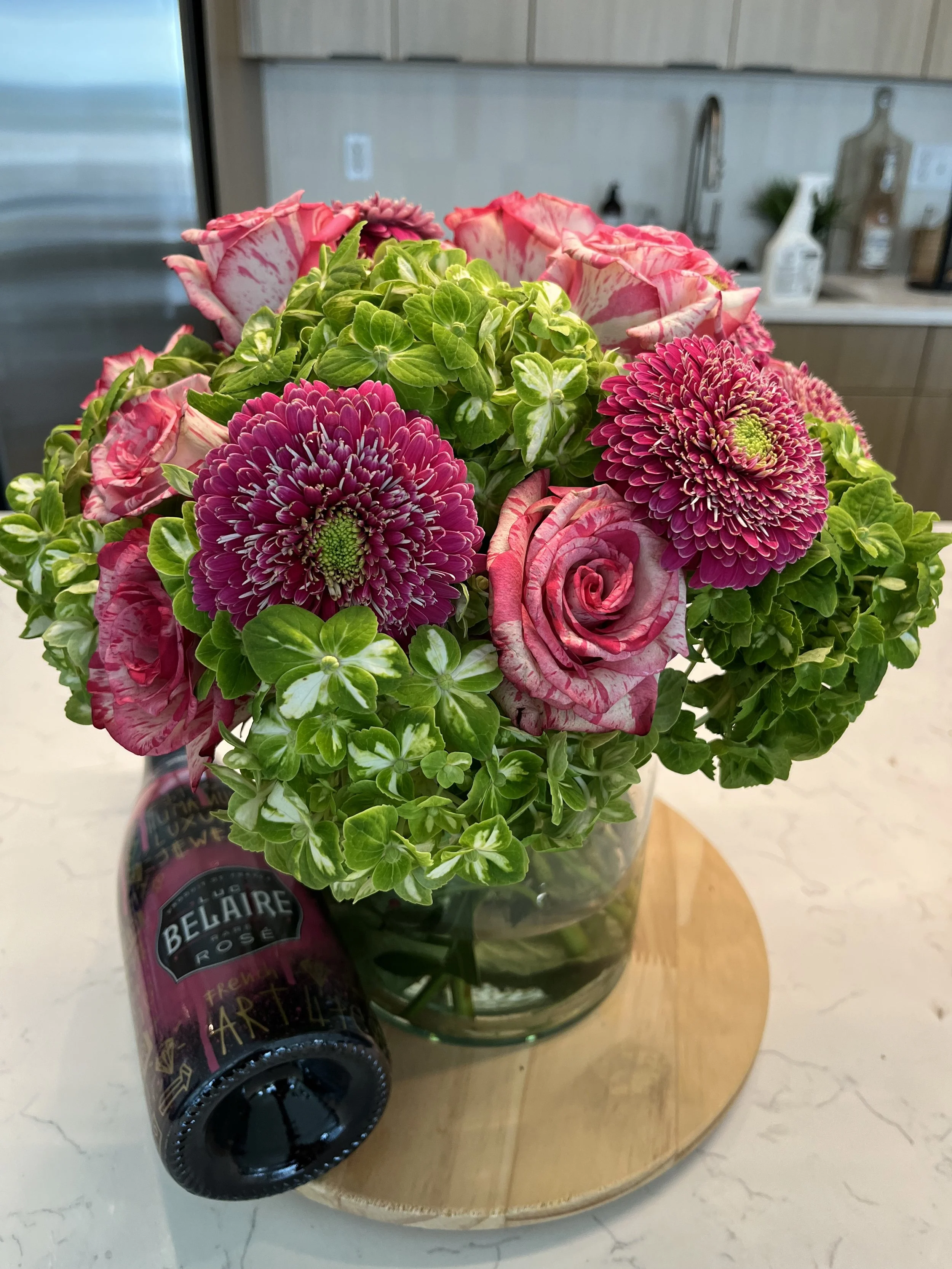 A bouquet of pink roses, green hydrangeas, and purple chrysanthemums in a glass vase on a round wooden tray, with a bottle of beer with a black cap in the foreground.