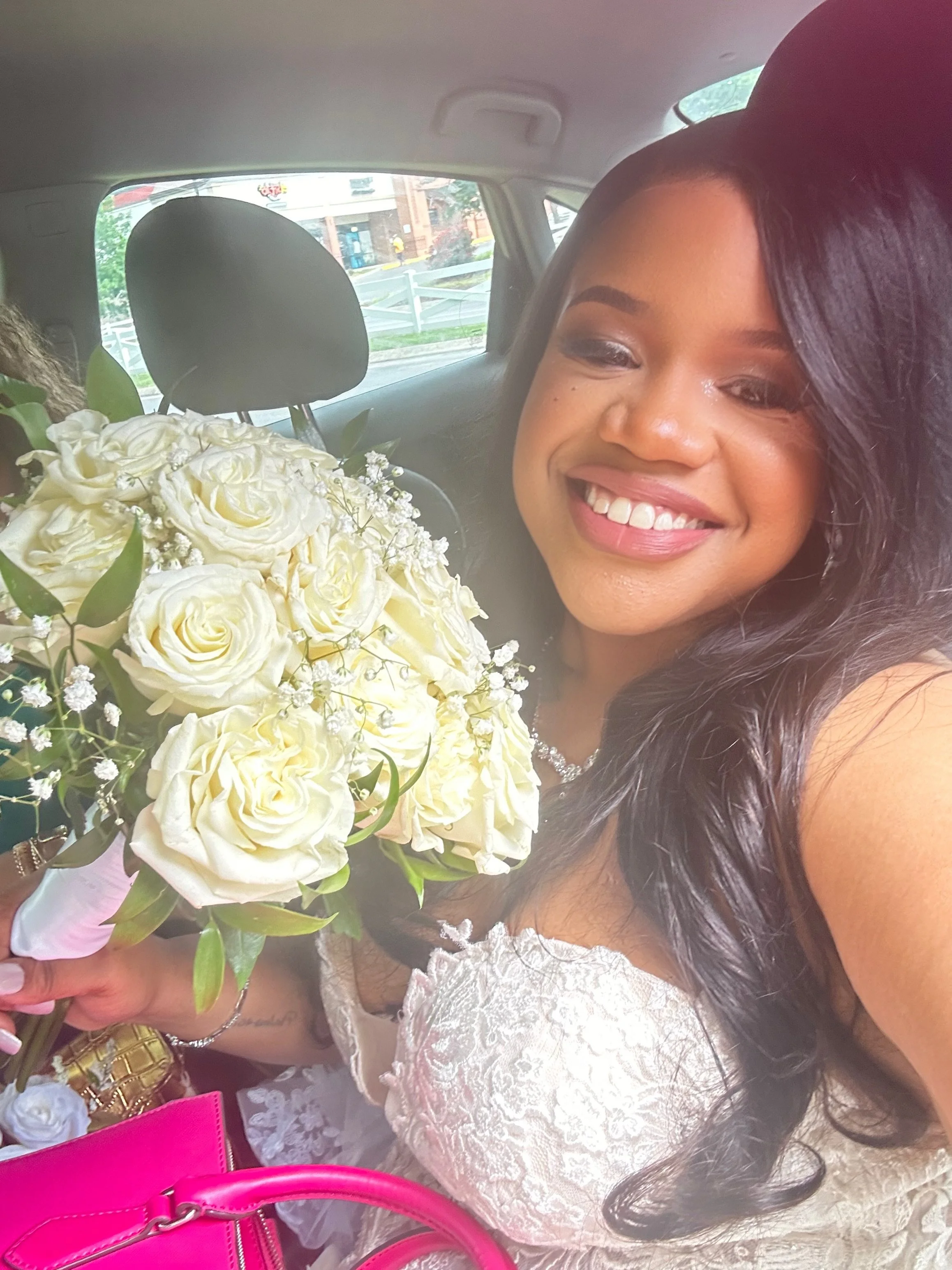A woman smiling, holding a large bouquet of white roses and greenery, dressed in white lace, sitting in a car.