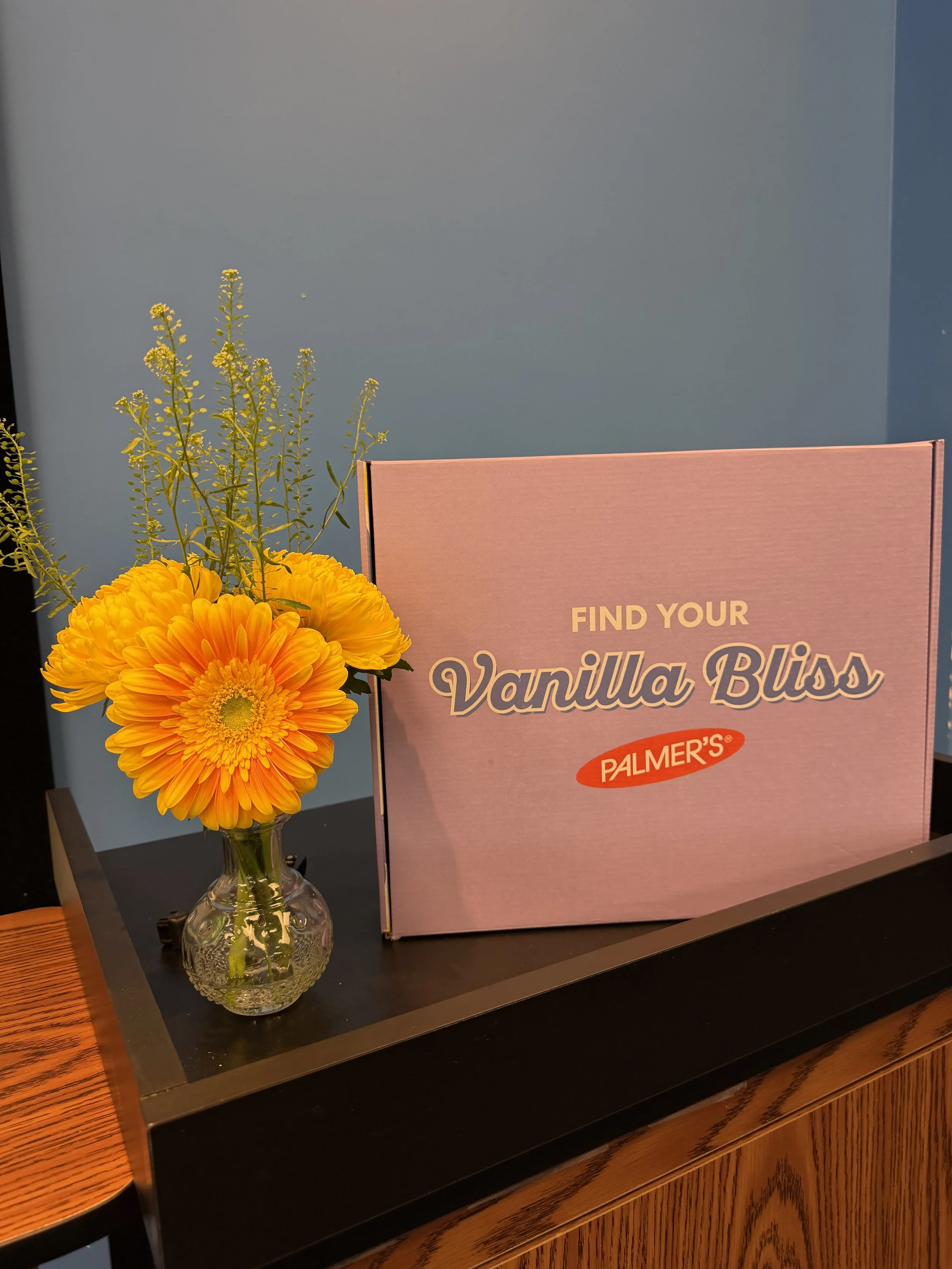 A small glass vase with orange and yellow flowers on a dark wooden table, next to a pink box with the text 'FIND YOUR Vanilla Bliss PALMER'S' on it.