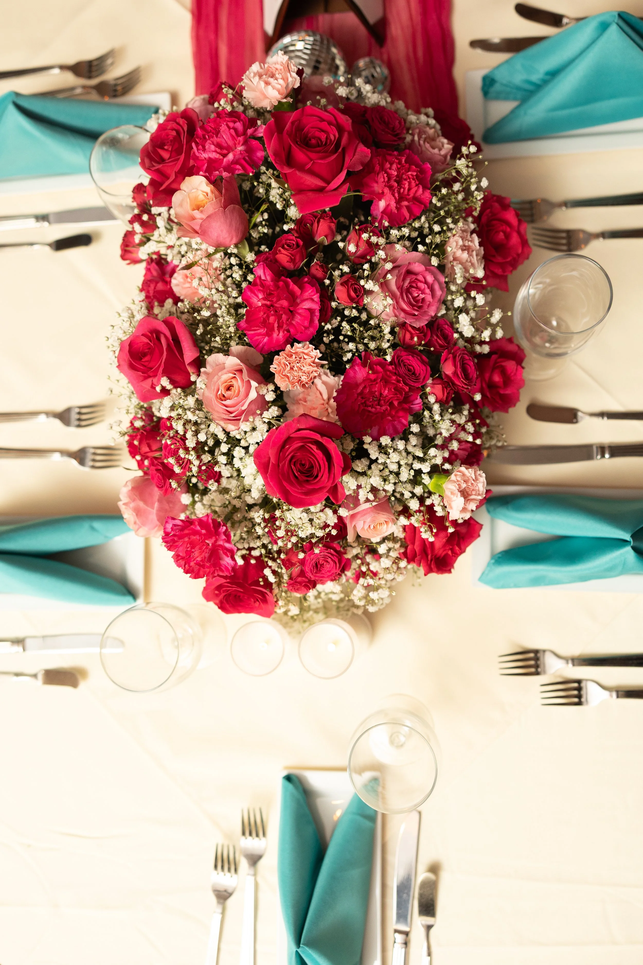 A beautifully arranged floral centerpiece with pink and red roses, pink carnations, and baby's breath on a dining table set for a meal with turquoise and white napkins and glassware.